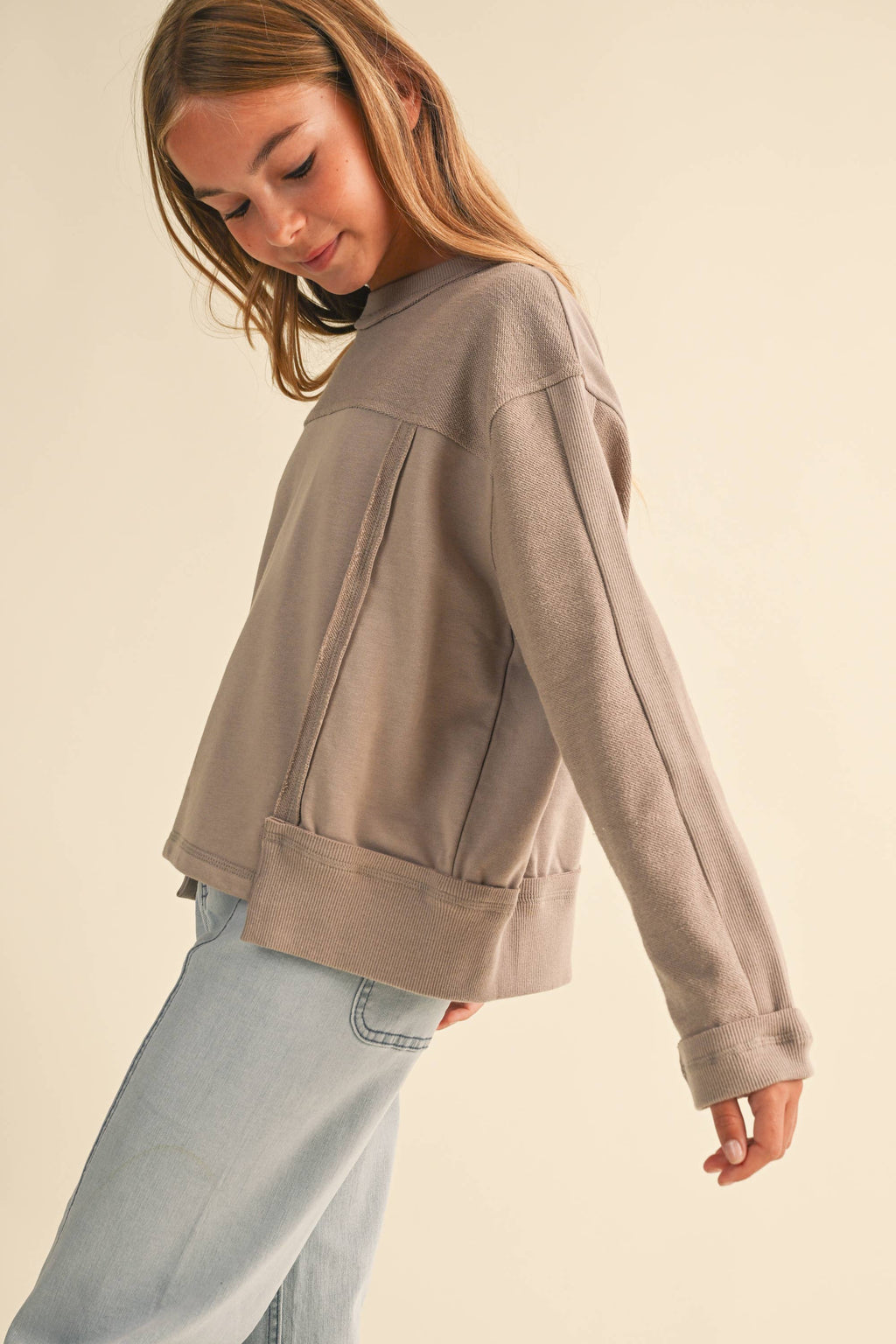 A young woman wearing a taupe-brown cropped long-sleeve sweatshirt and light-wash jeans, photographed against a solid cream background.