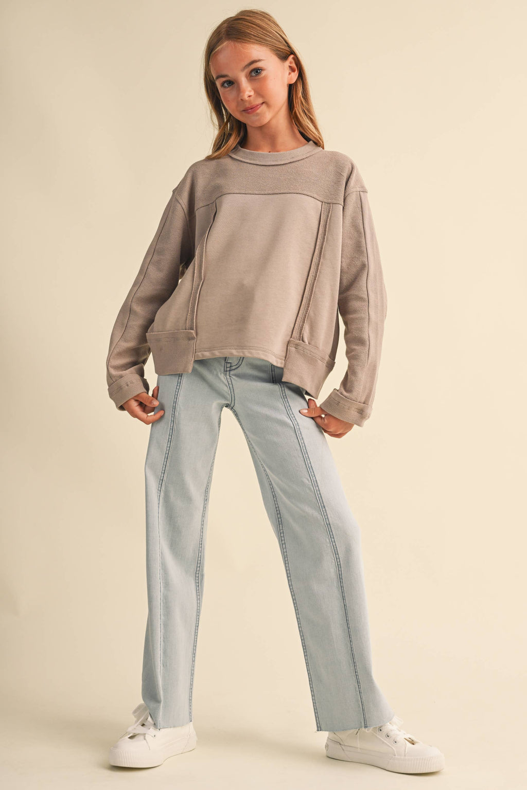 A young girl modeling a taupe-brown color-blocked sweatshirt and light-wash blue jeans against a solid beige studio background.