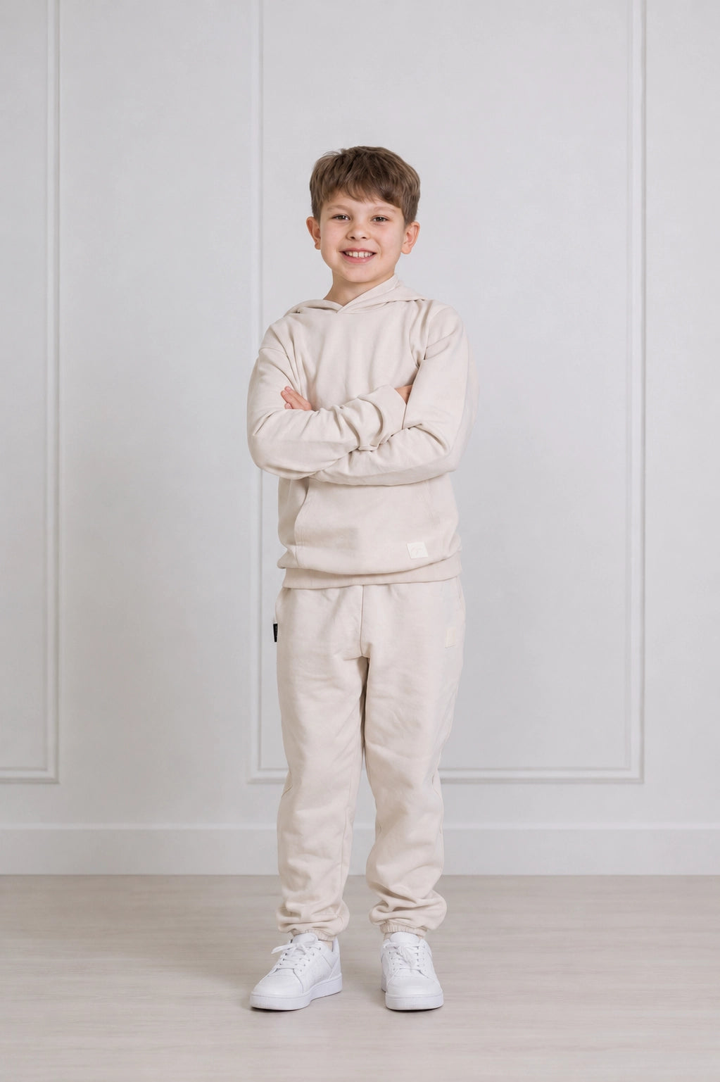 A young boy stands with arms crossed wearing a matching light beige hoodie and sweatpants set, paired with white sneakers, in front of a white paneled wall.