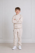 A young boy stands with arms crossed wearing a matching light beige hoodie and sweatpants set, paired with white sneakers, in front of a white paneled wall.