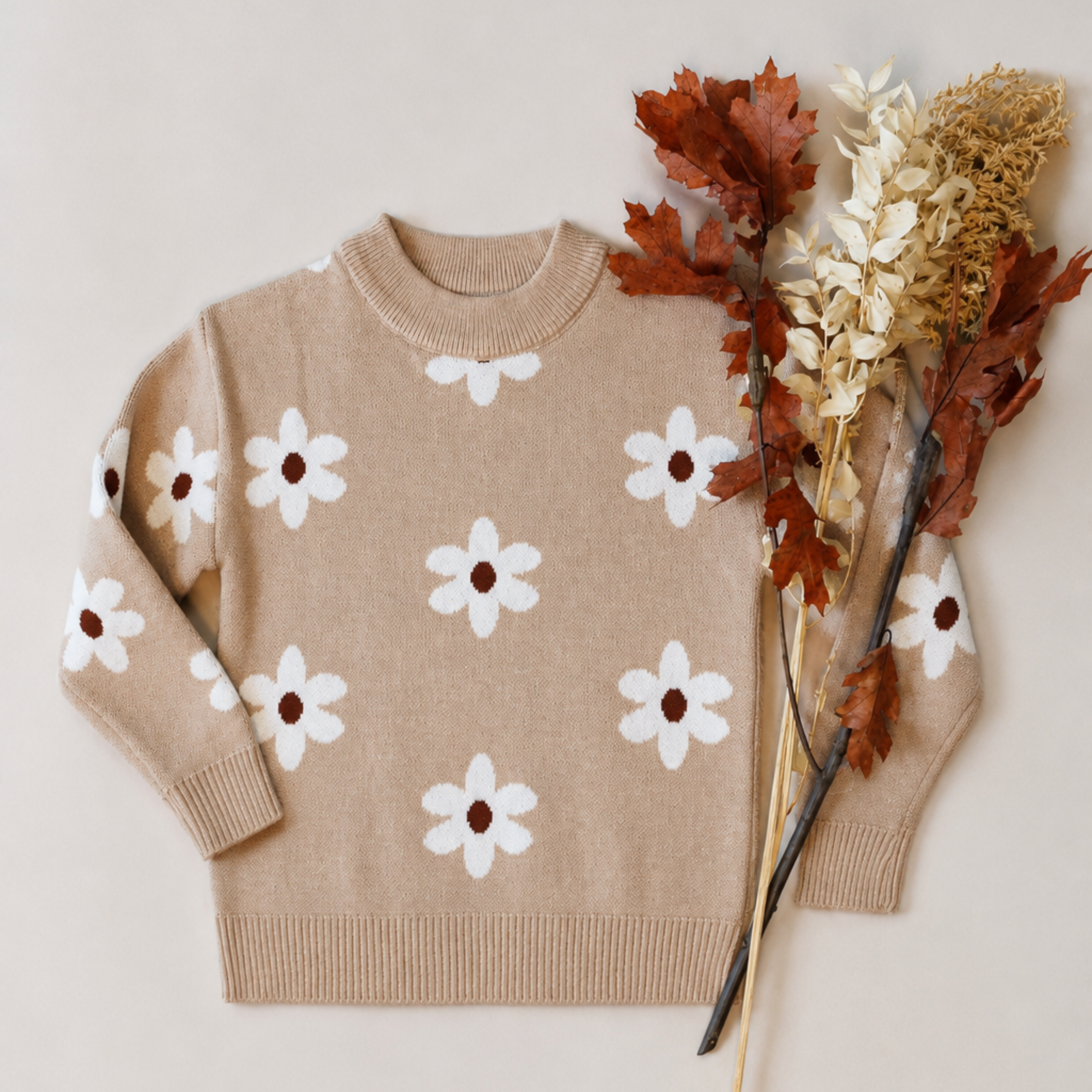 A beige knit sweater with a white daisy pattern, styled flat-lay with dried autumn flowers on a light beige background.