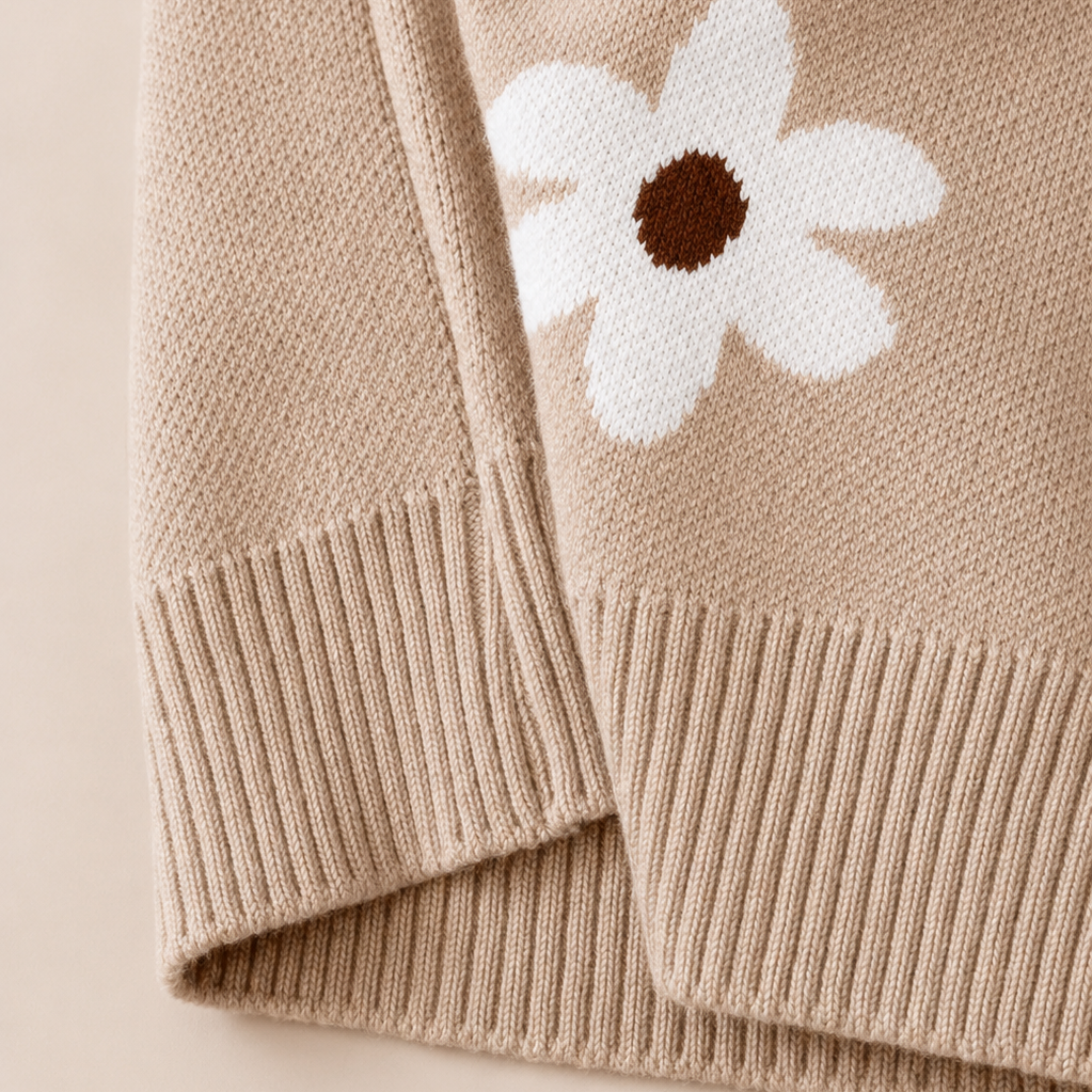 Close-up of a beige knit sweater with a white flower motif featuring a brown center, shown with ribbed cuffs and hem.