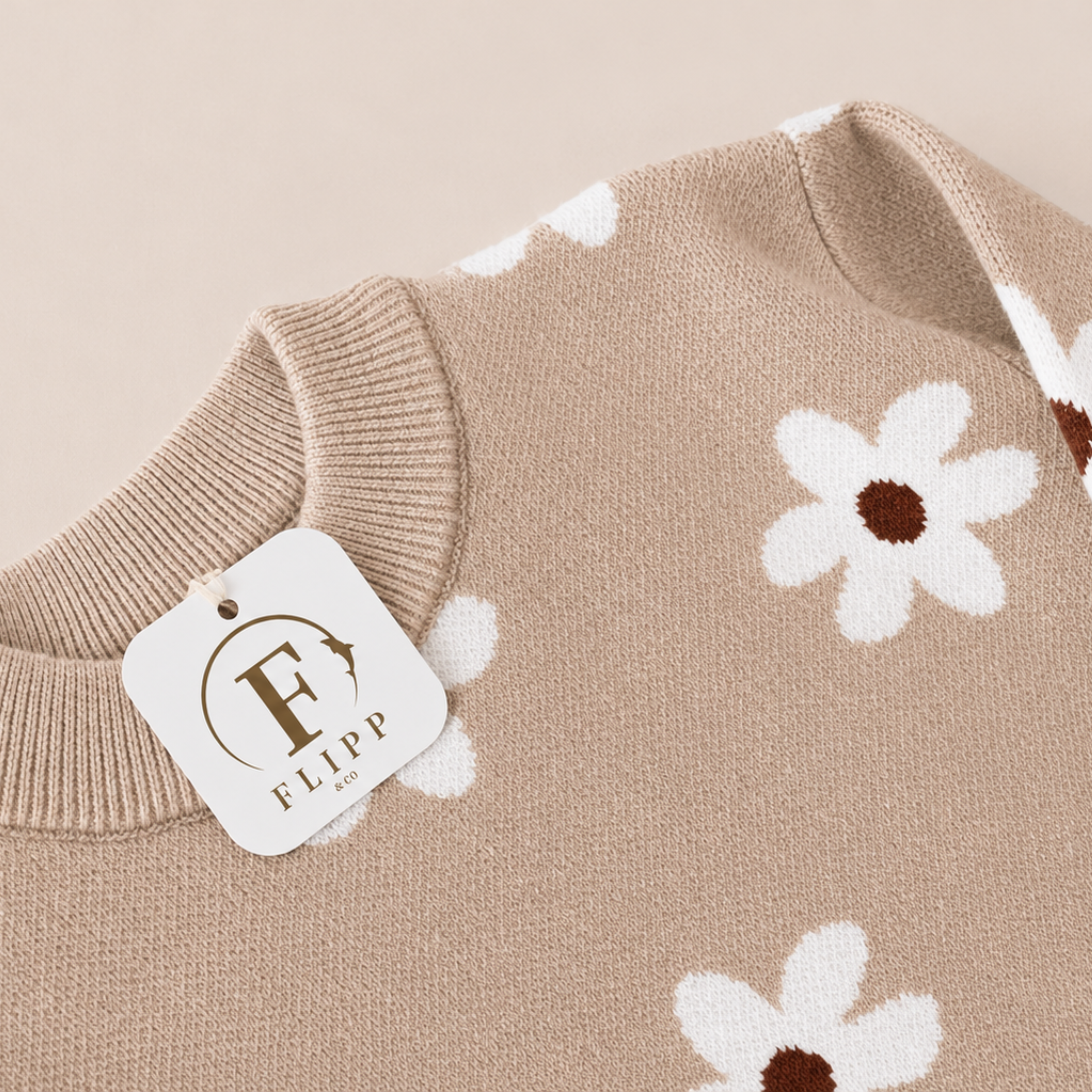 Close-up of a beige knit sweater with a white daisy pattern and a white hanging tag reading FLIPP.
