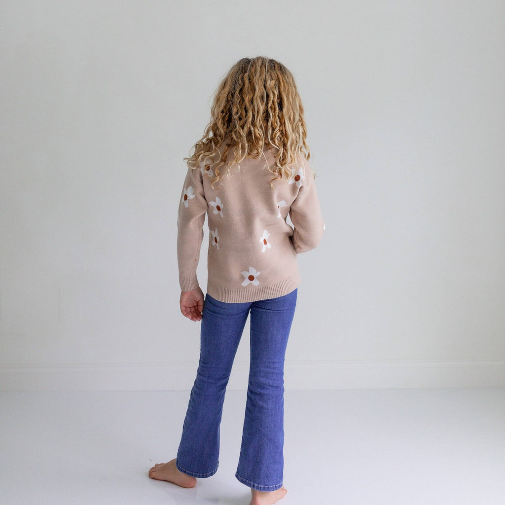 Child wearing a beige sweater with floral patterns and blue jeans on a plain background