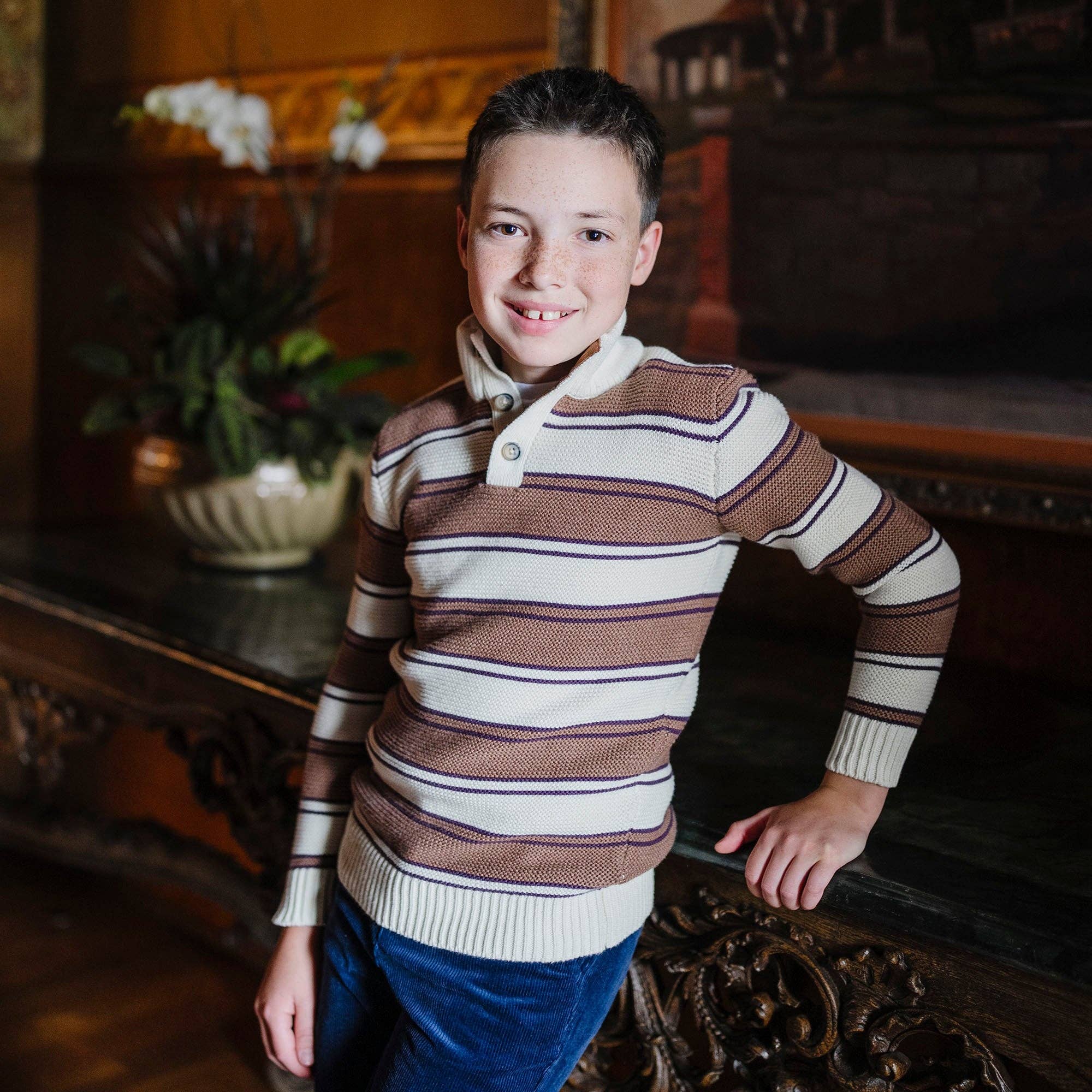 A young boy smiling while wearing a brown and cream striped knit polo sweater with blue jeans, standing in a warm, dark wood-paneled room.