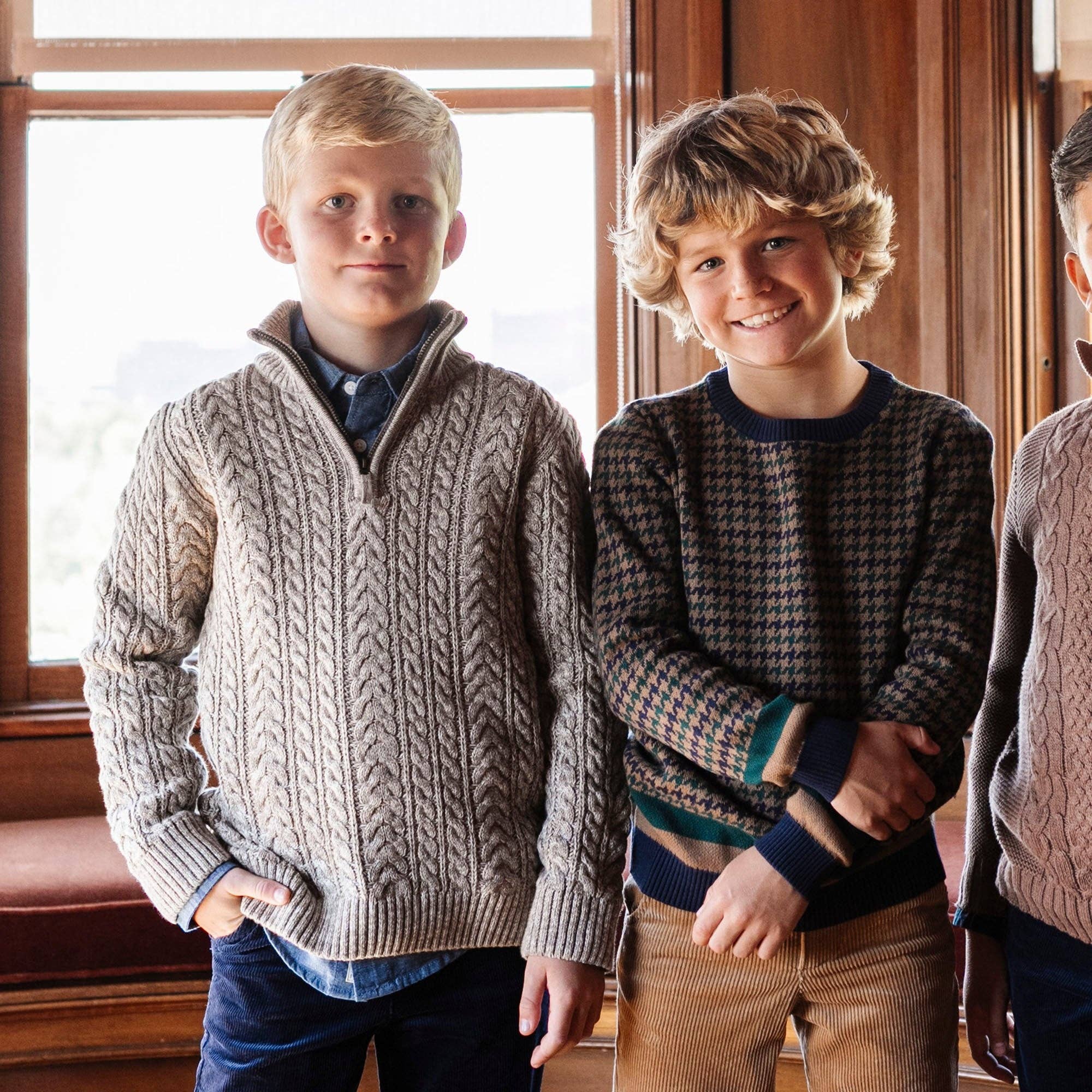 Two boys wearing knit sweaters in a warm indoor setting with wood paneling and a window. The boy on the left wears a beige cable-knit quarter-zip sweater, and the boy on the right wears a dark houndstooth patterned sweater.
