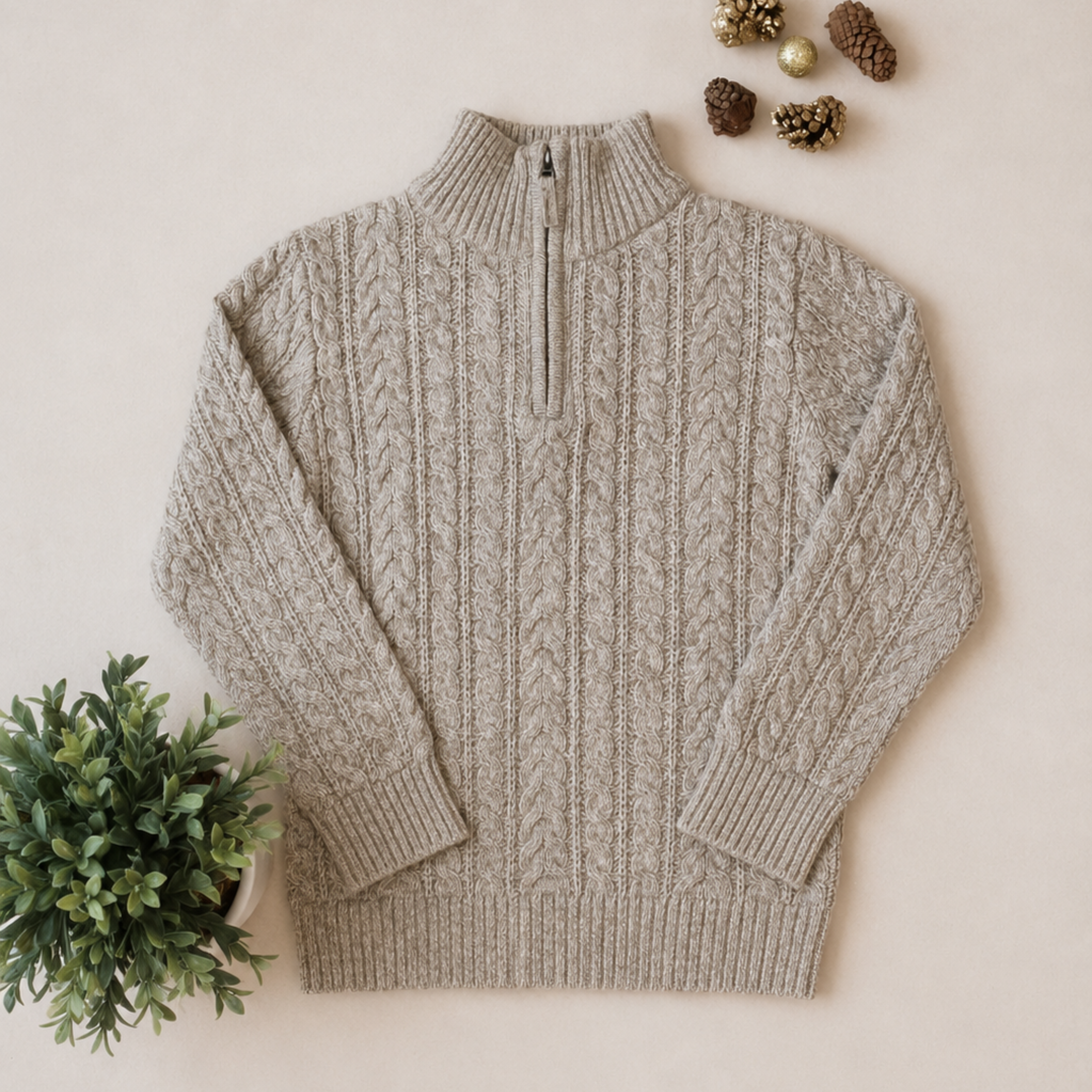 A beige cable-knit quarter-zip sweater with a mock neck, laid flat on a light beige background with decorative pinecones and a small green plant.