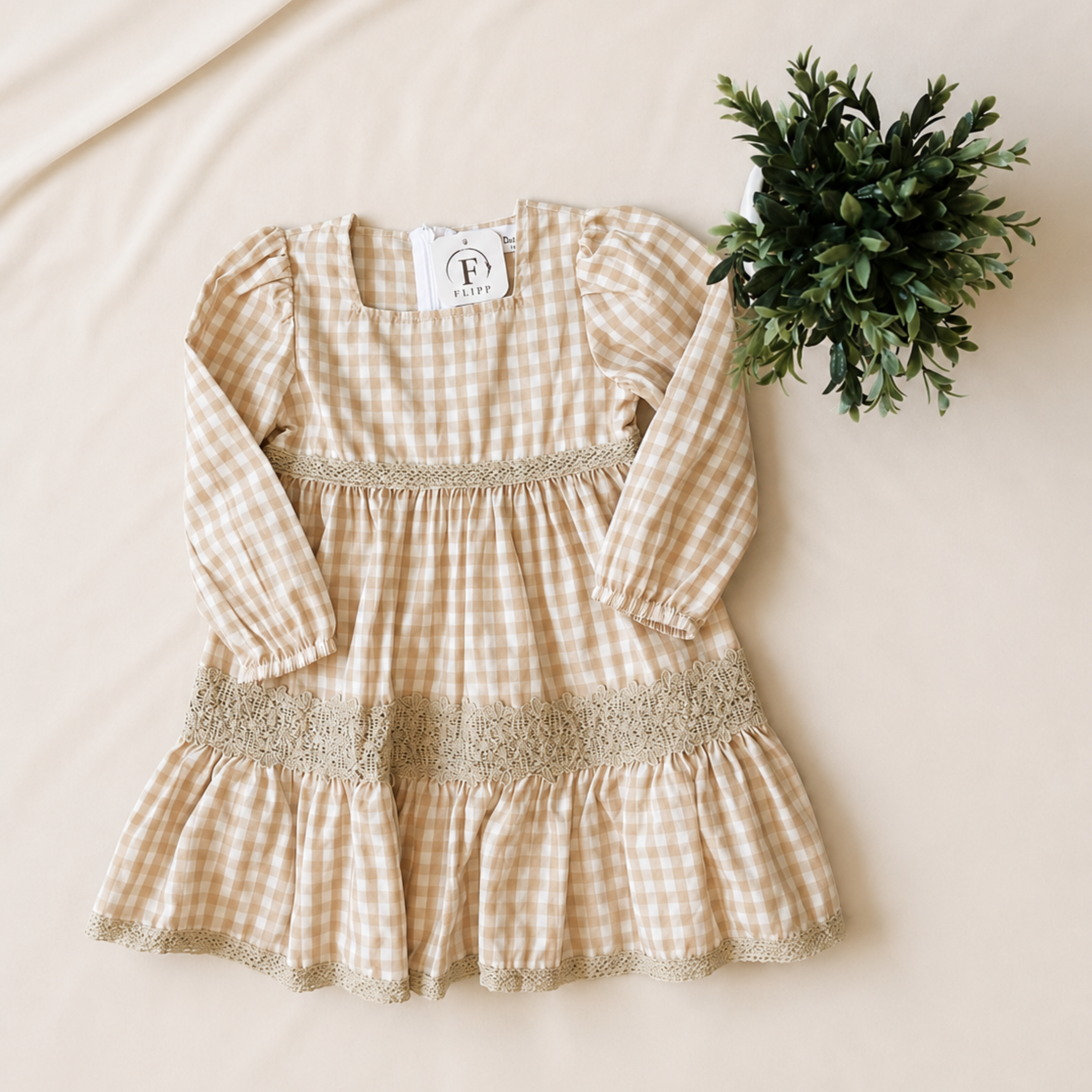 A toddler girl's long-sleeve dress in a beige and white gingham pattern with lace trim, laid flat on a beige fabric background next to a small green plant.