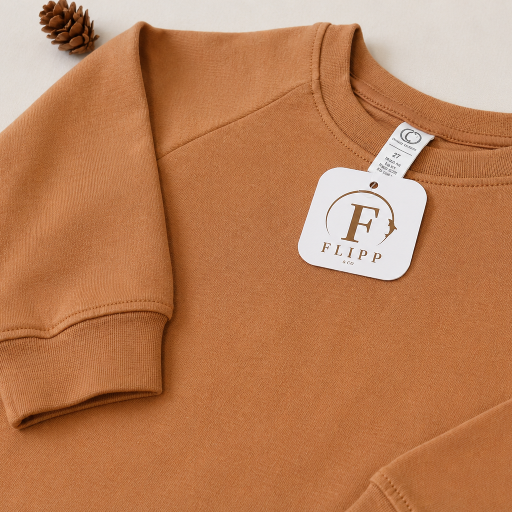 A close-up of a crewneck sweatshirt in a warm camel brown color, featuring a white brand tag for 'FLIPP' and a small pinecone prop on a light beige background.