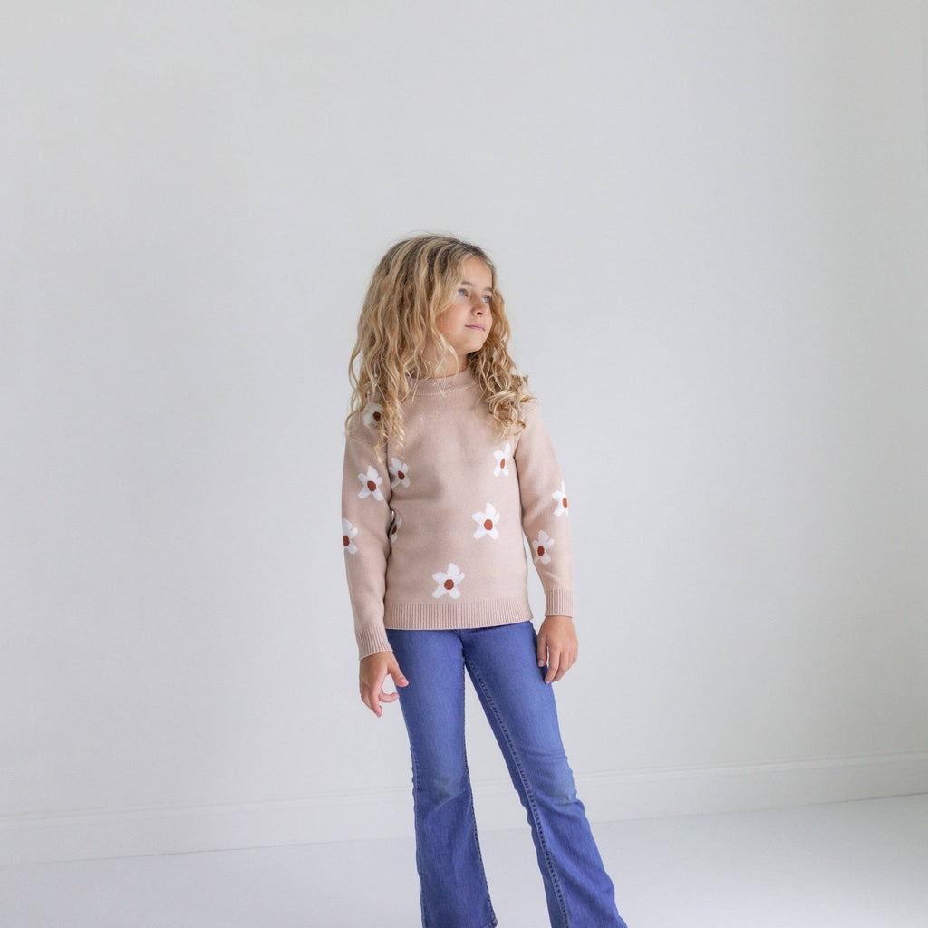 Person wearing a light brown sweater with floral patterns and blue jeans on a white background
