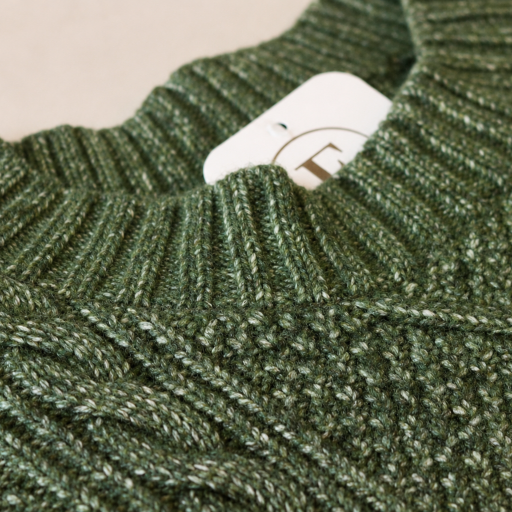 Close-up of a textured knit sweater in a heathered olive green color, showing the ribbed collar and a white paper tag with a brown logo.