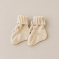 A pair of cream-colored hand-knit baby booties with ribbed cuffs and small tie details, displayed on a neutral beige background.