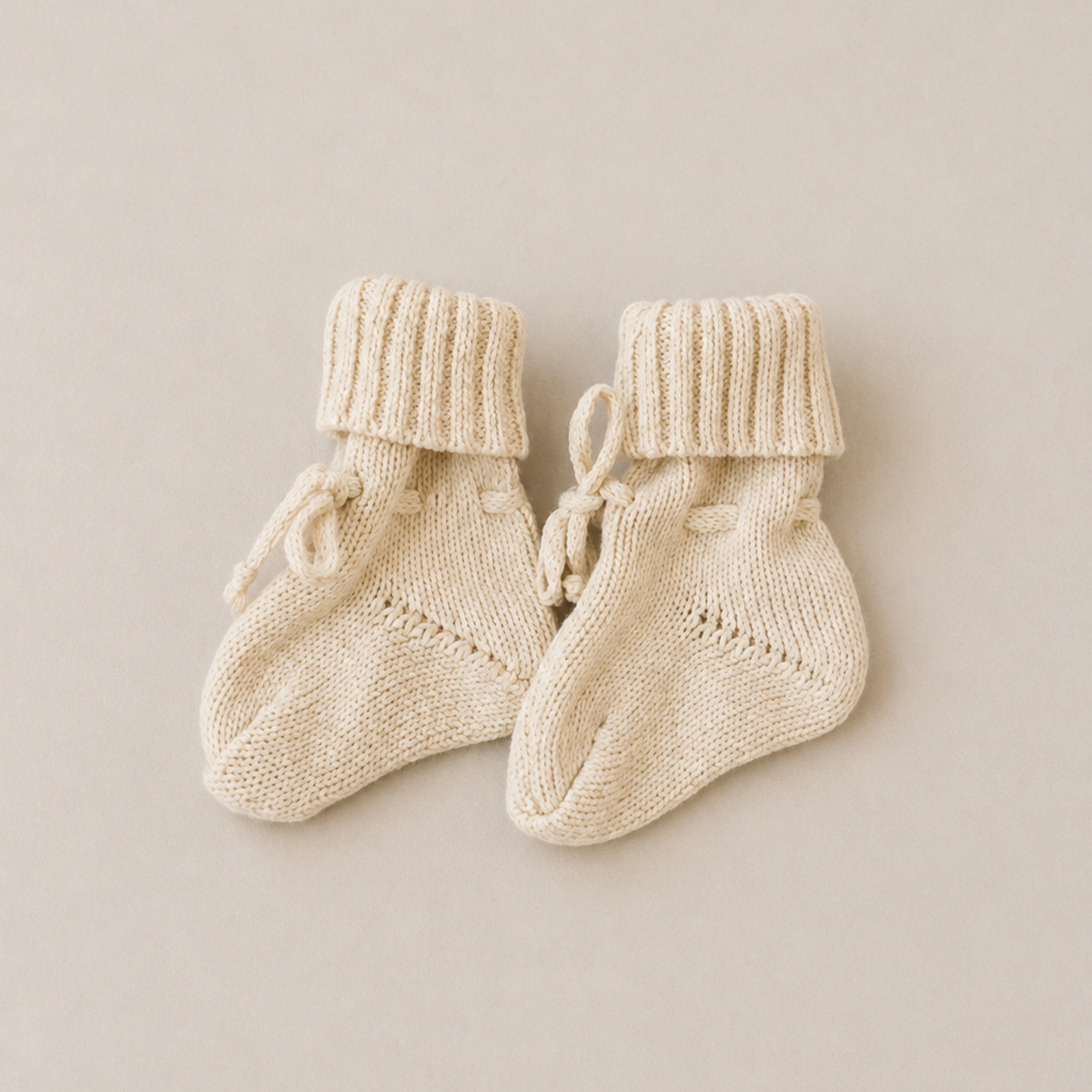 A pair of cream-colored hand-knit baby booties with ribbed cuffs and small tie details, displayed on a neutral beige background.