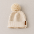 A cream-colored knit beanie with a matching pom-pom and a small brown leather brand tag, photographed flat on a neutral beige background.