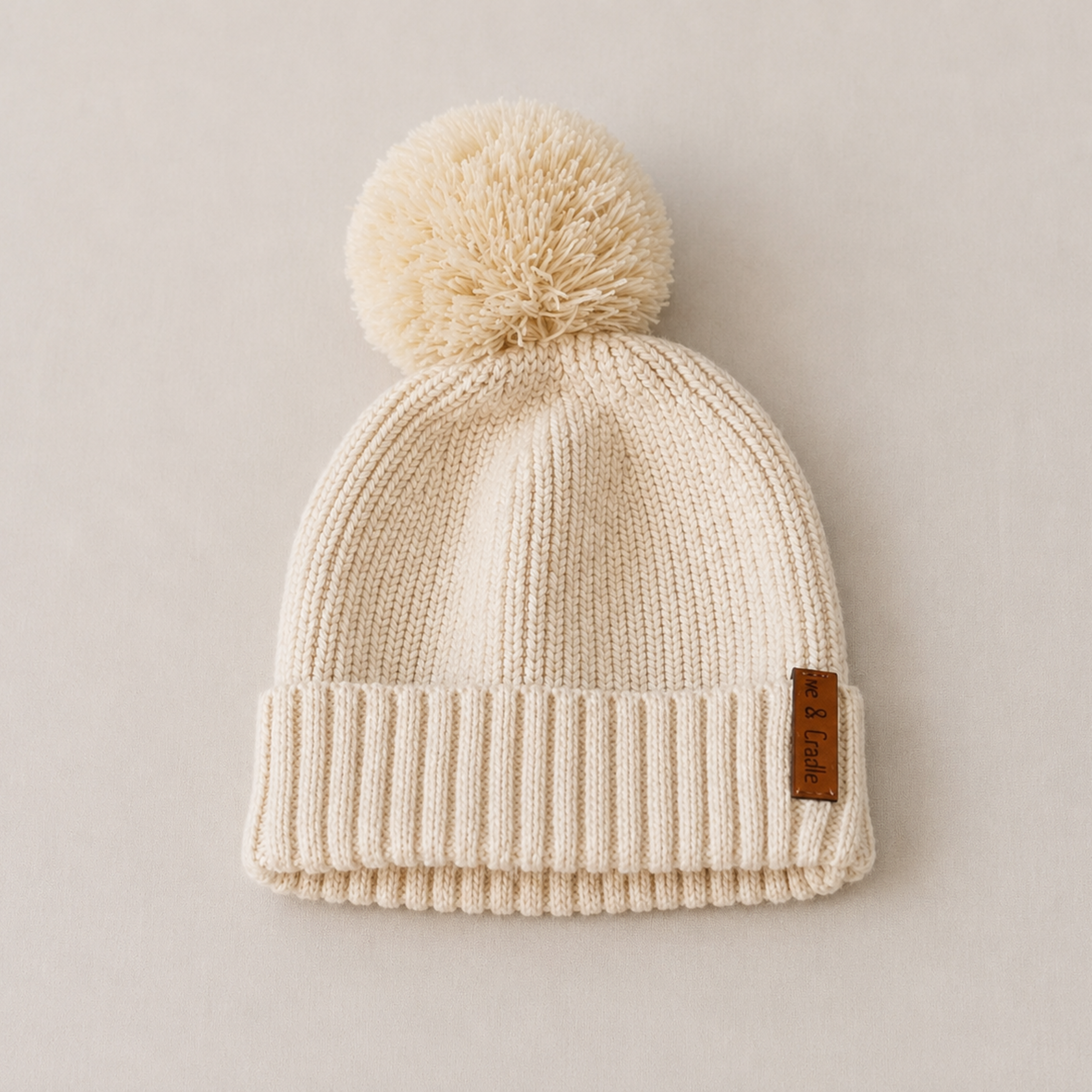 A cream-colored knit beanie with a matching pom-pom and a small brown leather brand tag, photographed flat on a neutral beige background.