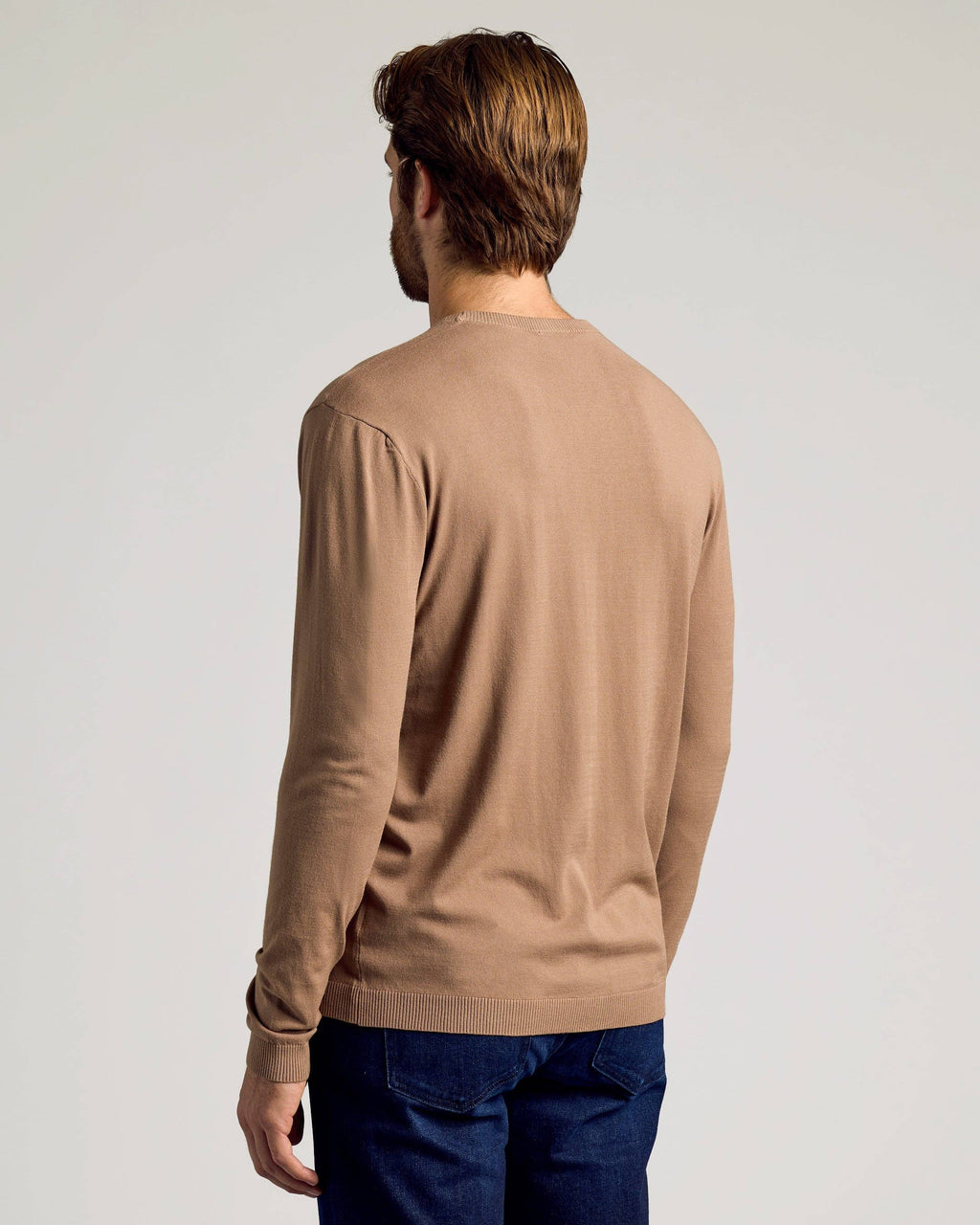 A man viewed from the back wearing a light brown long-sleeve knit sweater and dark blue jeans, set against a plain light gray studio background.