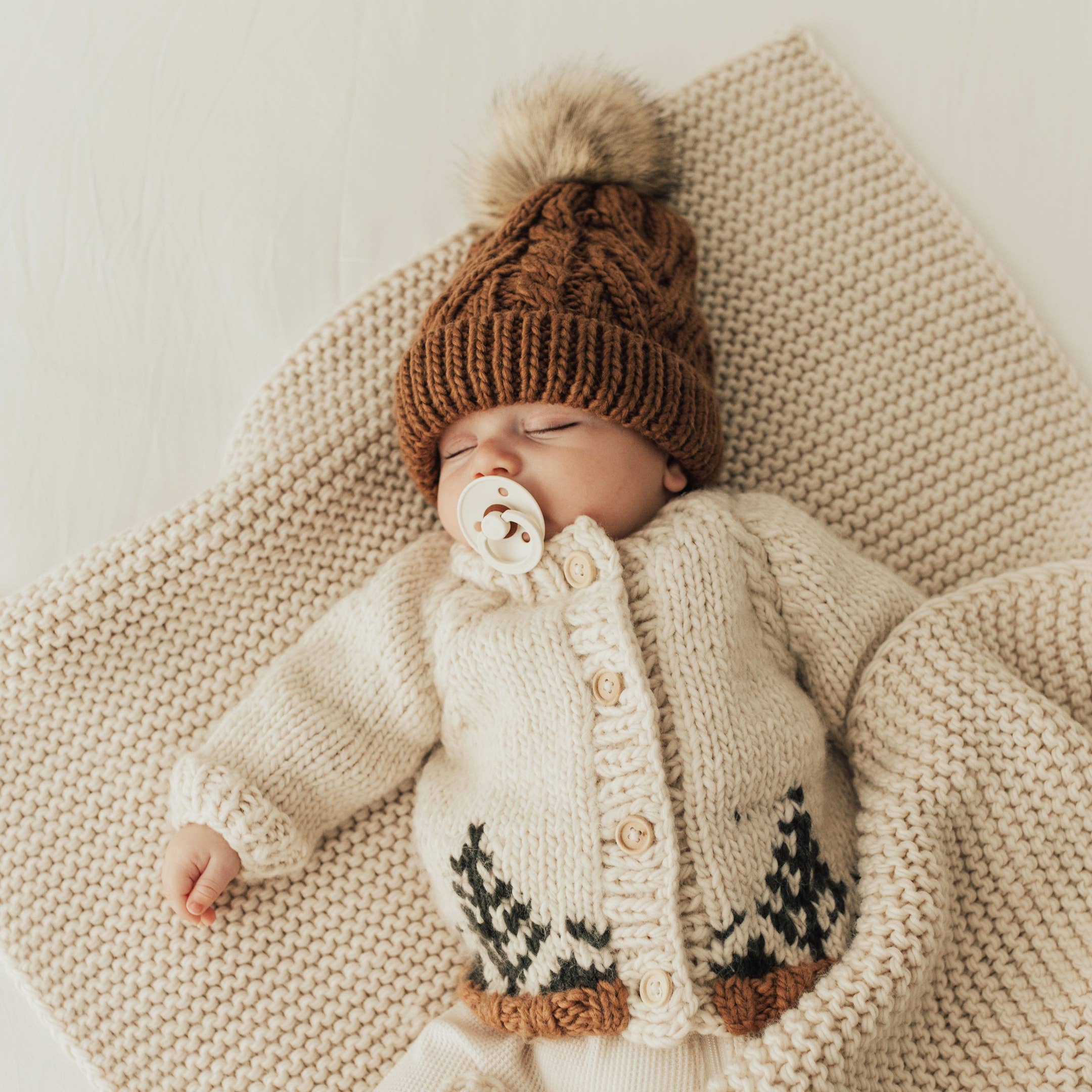 Baby wearing a knitted cardigan and hat with a pom-pom, lying on a textured blanket.