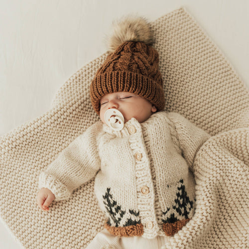 Baby wearing a knitted cardigan and hat with a pom-pom, lying on a textured blanket.