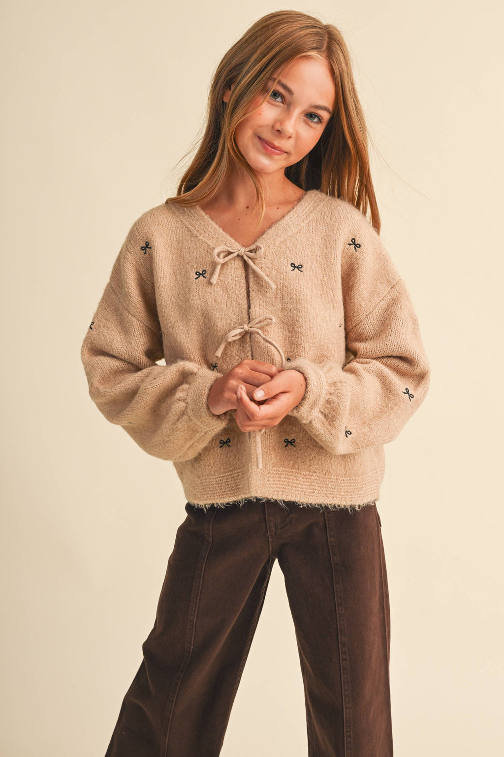 A young girl with long light brown hair wearing a beige fuzzy knit sweater with butterfly details and a tie neckline, paired with dark brown pants, against a solid cream background.