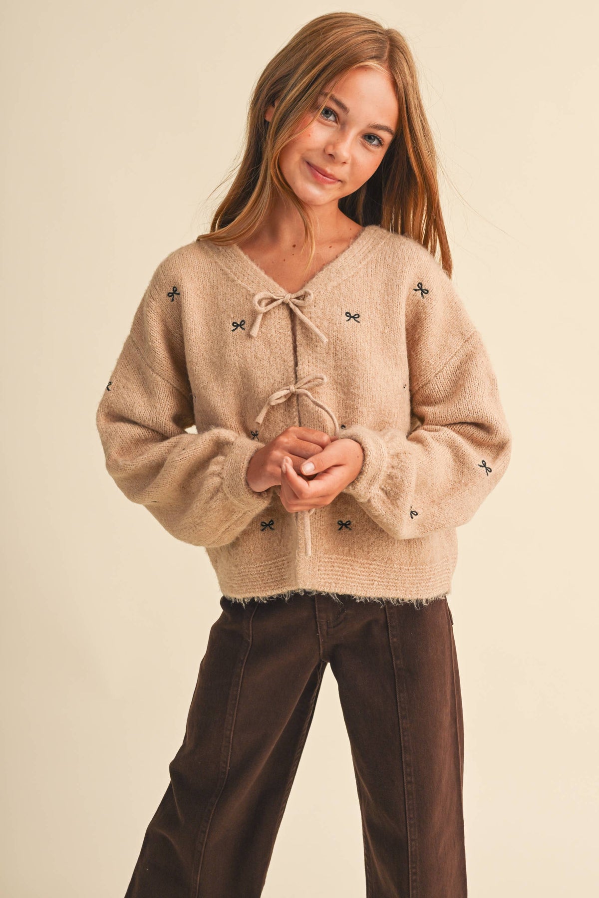 A young girl with long light brown hair wearing a beige fuzzy knit sweater with butterfly details and a tie neckline, paired with dark brown pants, against a solid cream background.