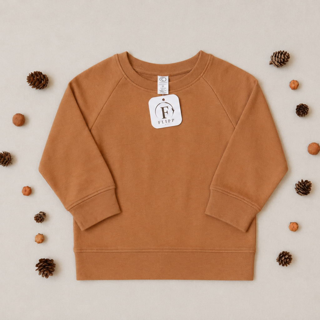 A child's long-sleeve crewneck sweatshirt in a warm camel brown color, laid flat on a light beige background and surrounded by decorative pinecones and small orange fruits. A white brand tag for 'Fetty' is attached to the collar.