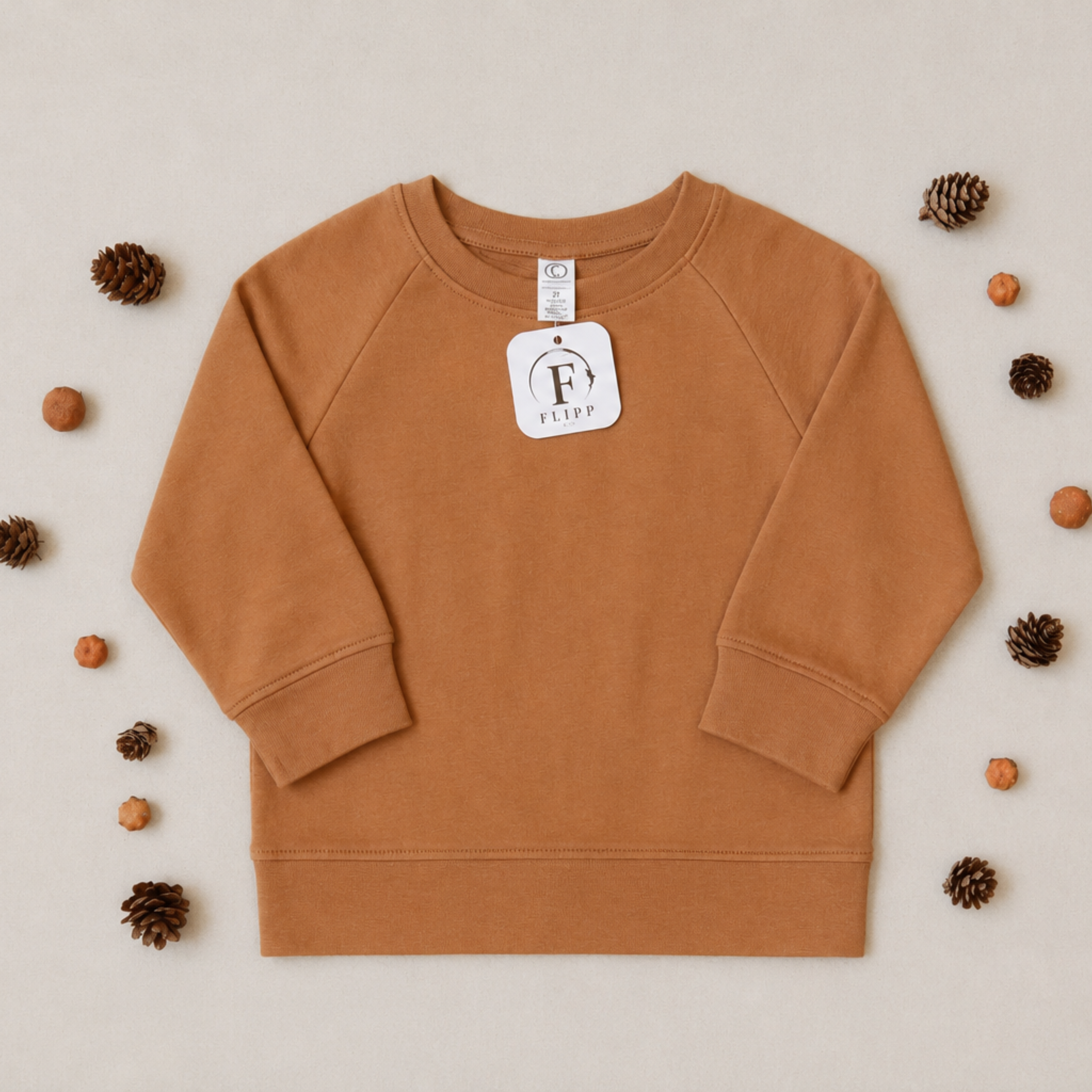 A child's long-sleeve crewneck sweatshirt in a warm camel brown color, laid flat on a light beige background and surrounded by decorative pinecones and small orange fruits. A white brand tag for 'Fetty' is attached to the collar.