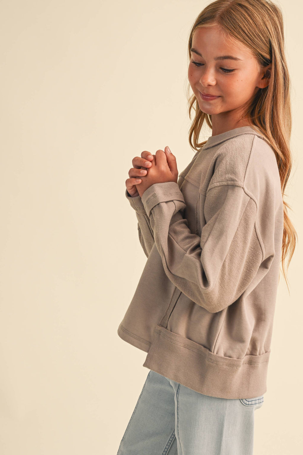 A young girl with long light brown hair is wearing a taupe-beige long-sleeve cropped sweatshirt and light blue jeans, standing against a solid cream background.