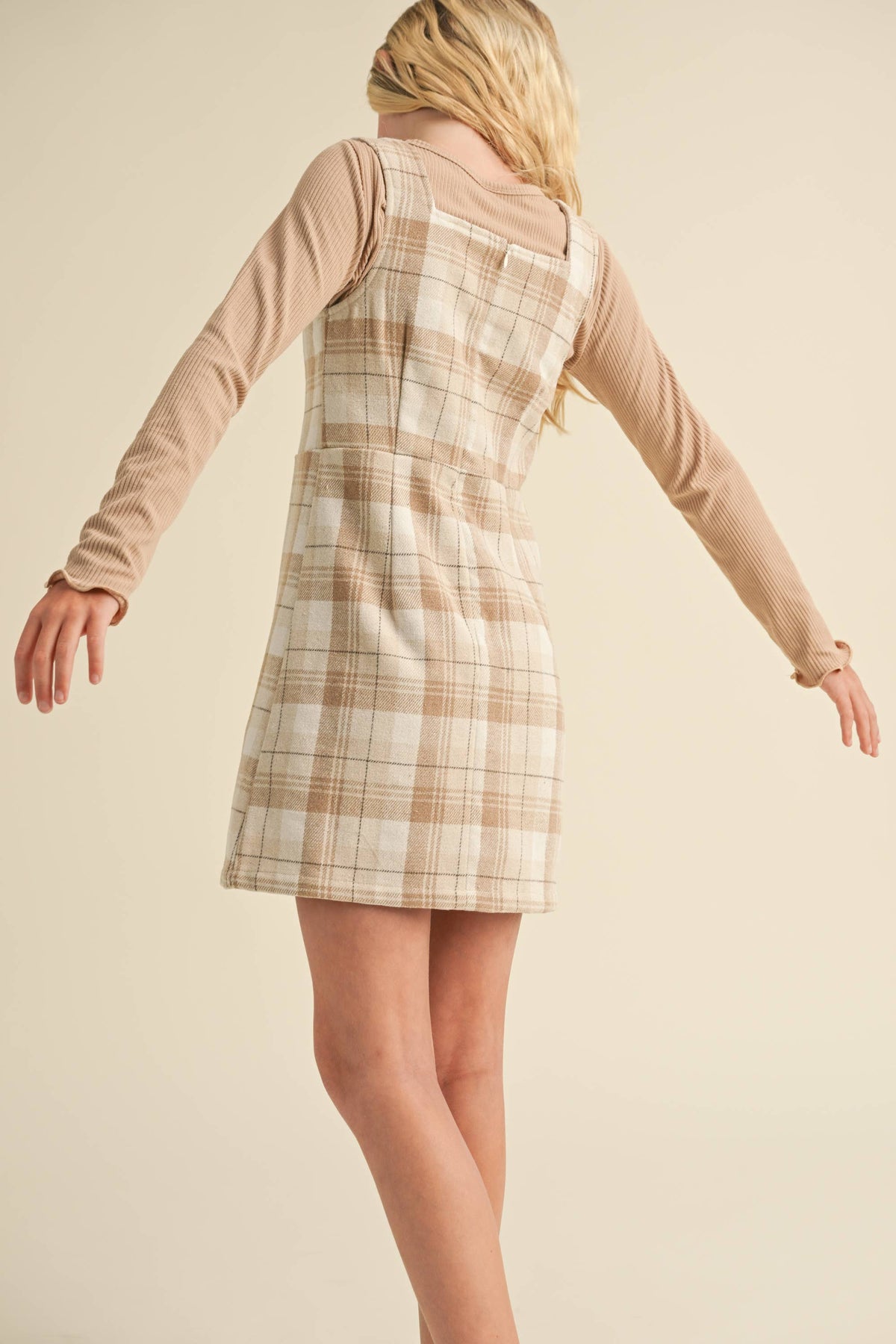 Person wearing a plaid dress over a long-sleeve shirt against a beige background