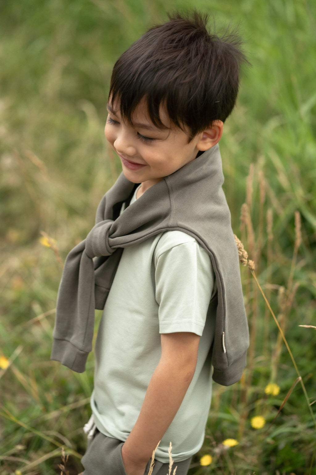 A young boy wearing a light sage green t-shirt and gray sweatpants, with a gray knit wrap or scarf tied around his neck, standing in a grassy outdoor field.