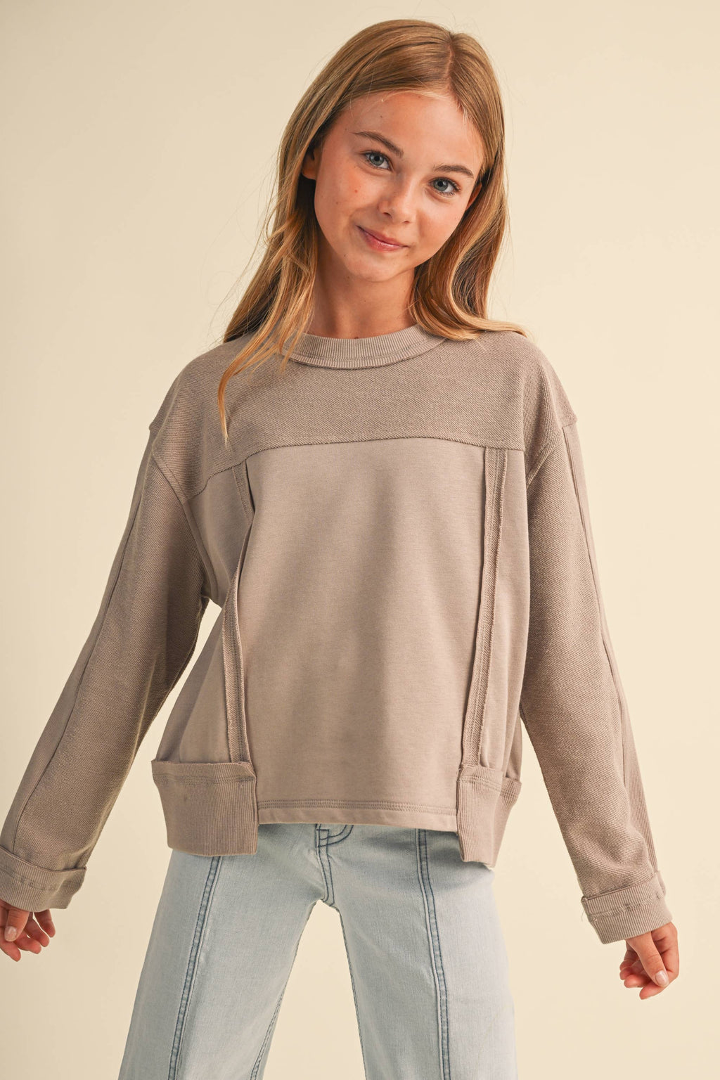 A young girl with blonde hair wearing a taupe long-sleeve crewneck sweatshirt and light blue jeans, standing against a solid beige background.