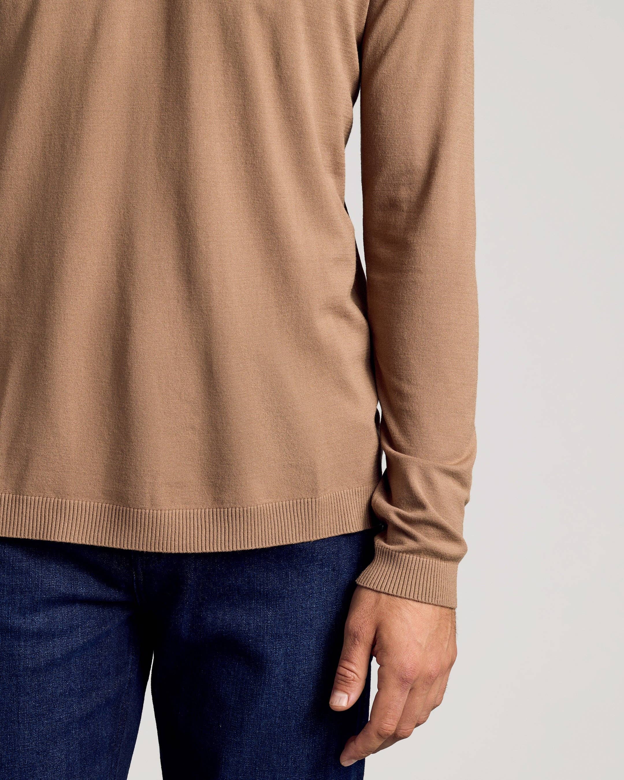A close-up of a person wearing a tan long-sleeve knit sweater with ribbed cuffs and hem, paired with dark blue jeans, against a light gray background.