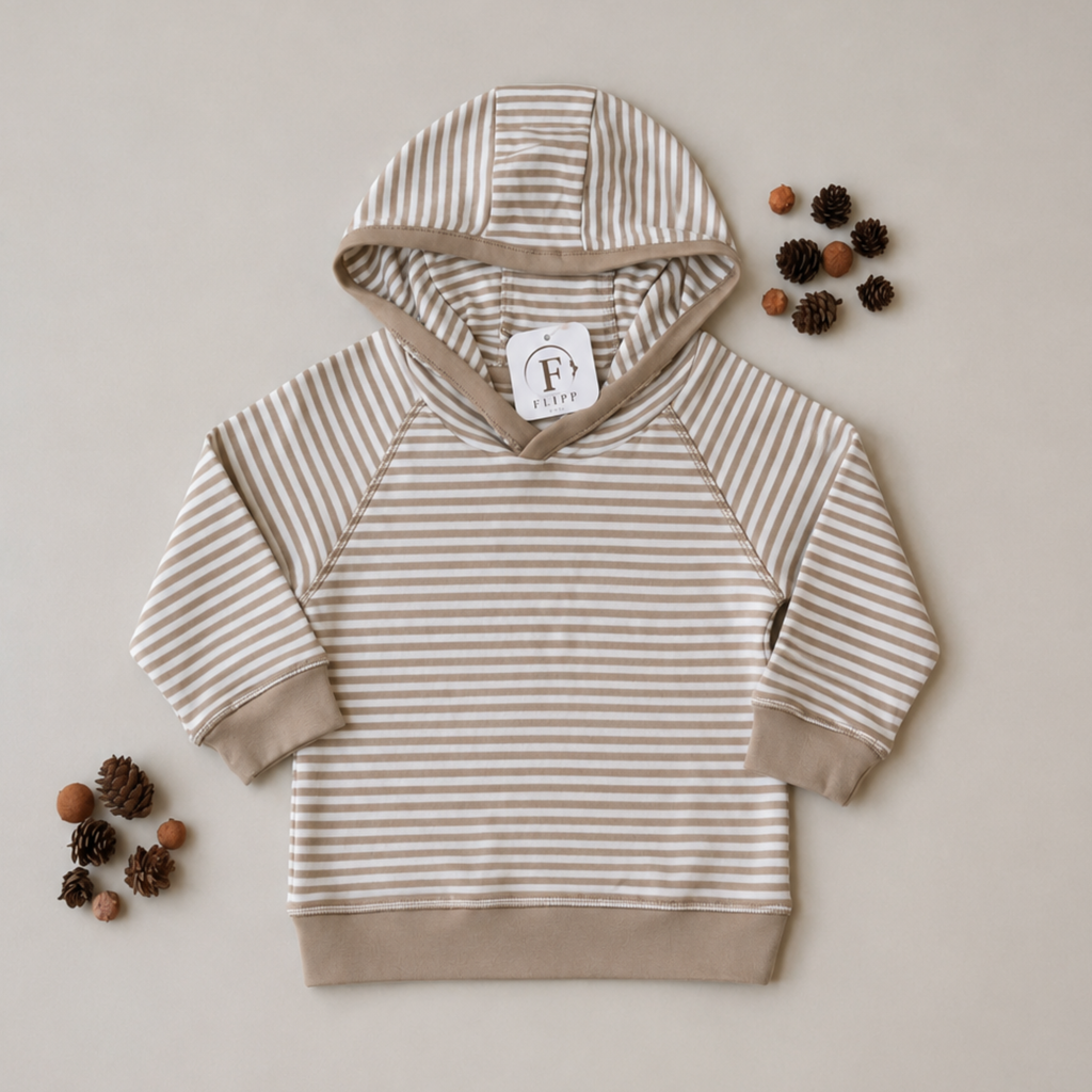 A children's long-sleeve hoodie with beige and white horizontal stripes, laid flat on a light beige background with decorative pinecones around it. The brand tag reads 'F+ Fille'.