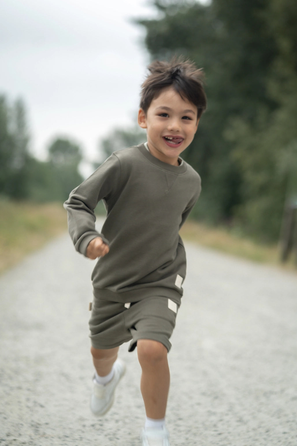 A young boy smiling and running on a gravel path outdoors, wearing an olive green long-sleeve sweatshirt and matching shorts with white sneakers.