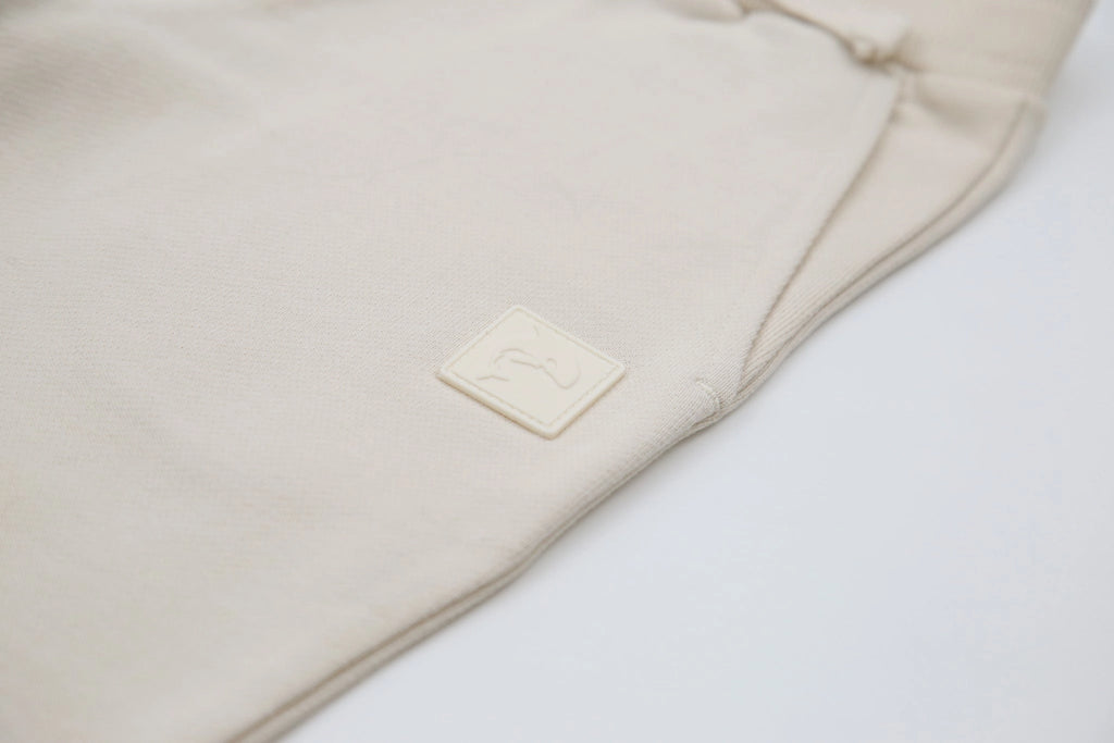 A close-up of a light beige fabric garment, likely sweatpants, featuring a small square embossed brand patch on the thigh.