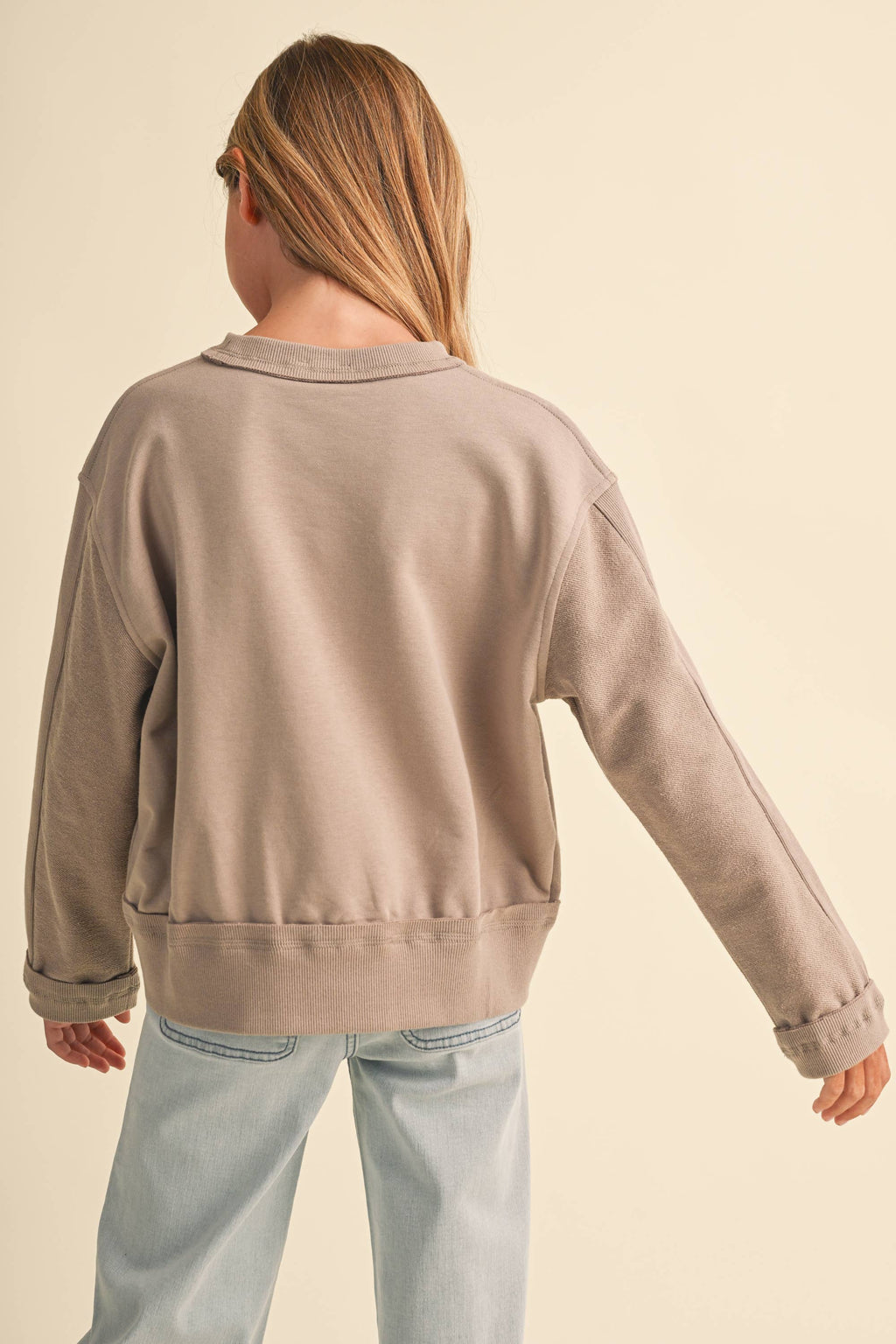 A child model shown from the back wearing a light taupe-beige crewneck sweatshirt and light blue jeans, set against a solid cream background.