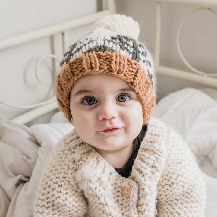 Baby wearing a knitted hat and coat sitting on a bed.