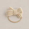 A cream-colored knitted bow hair tie with a matching beige elastic band, displayed on a neutral beige background.