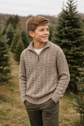 A young boy with light brown hair stands in a Christmas tree farm wearing a textured cable-knit quarter-zip sweater in taupe, dark olive pants, and a white t-shirt. The background consists of rows of green Christmas trees under an overcast sky.