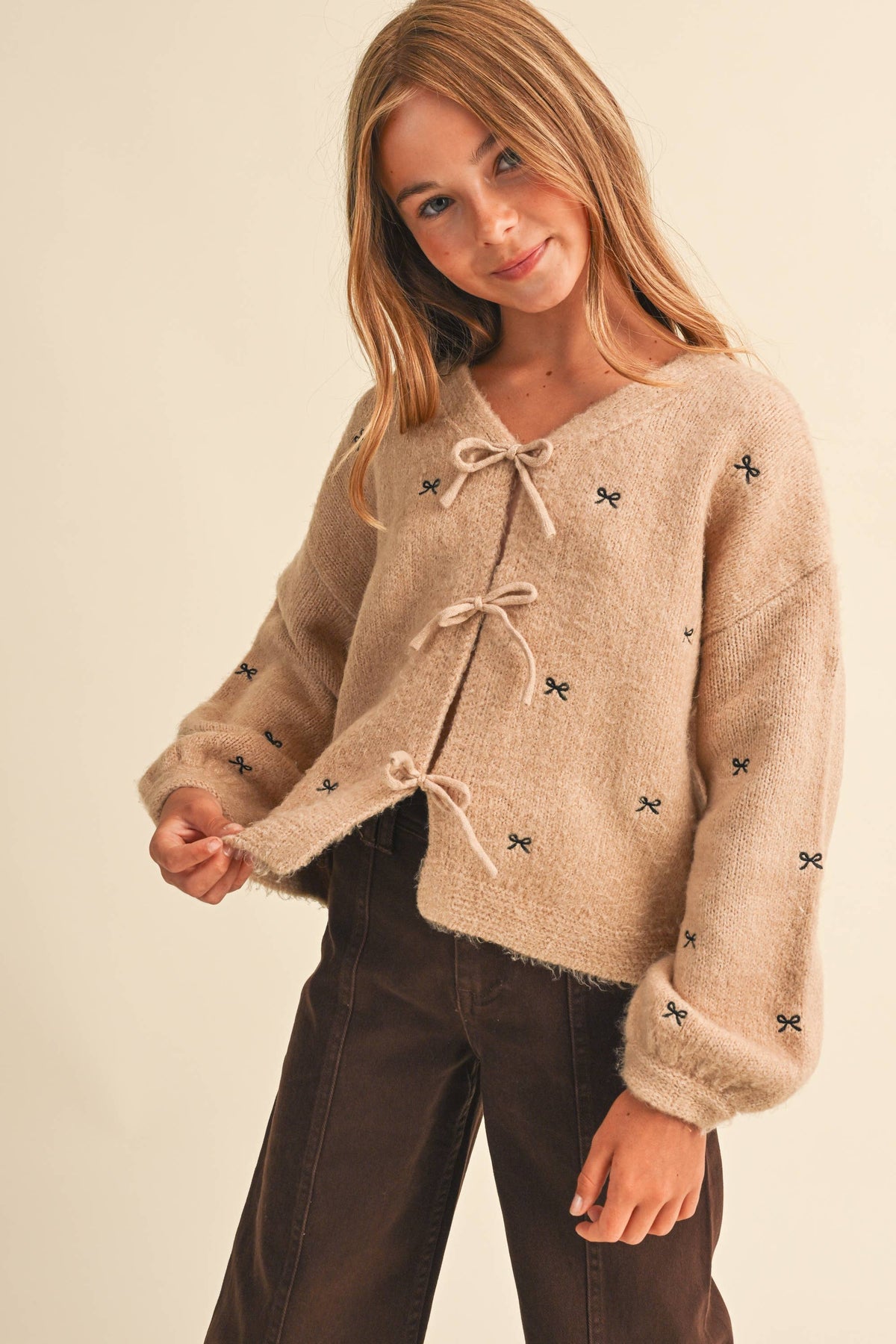 A young girl with blonde hair wearing a fuzzy beige cropped cardigan with tie closures and small black embroidered bow details, paired with dark brown pants, against a solid light beige background.