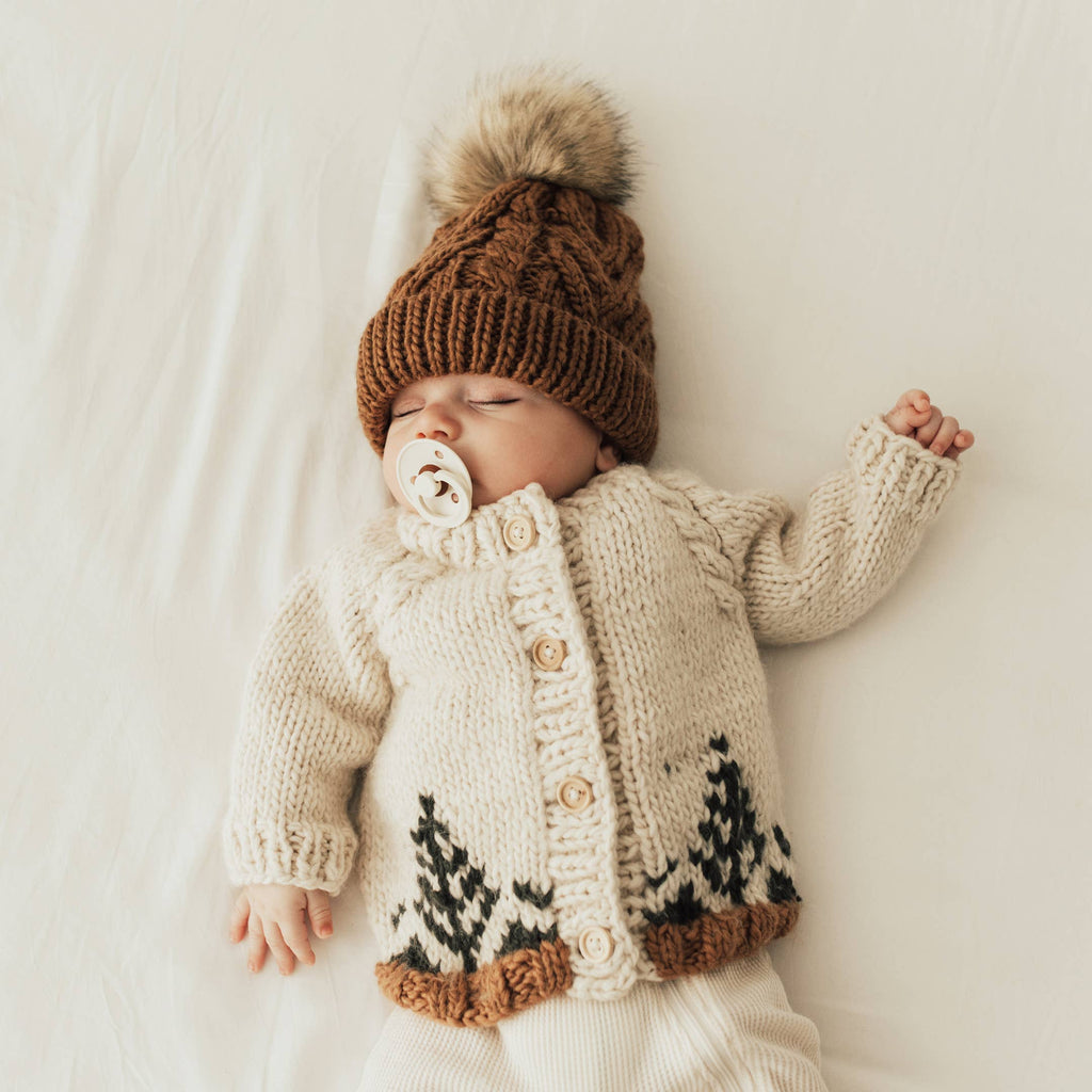 Baby wearing a knitted cardigan and hat with a pom-pom, lying on a white blanket.