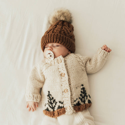 Baby wearing a knitted cardigan and hat with a pom-pom, lying on a white blanket.