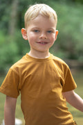 A young blond boy smiling at the camera, wearing a short-sleeve crewneck t-shirt in a warm mustard yellow color, set against a soft, blurred green outdoor background.