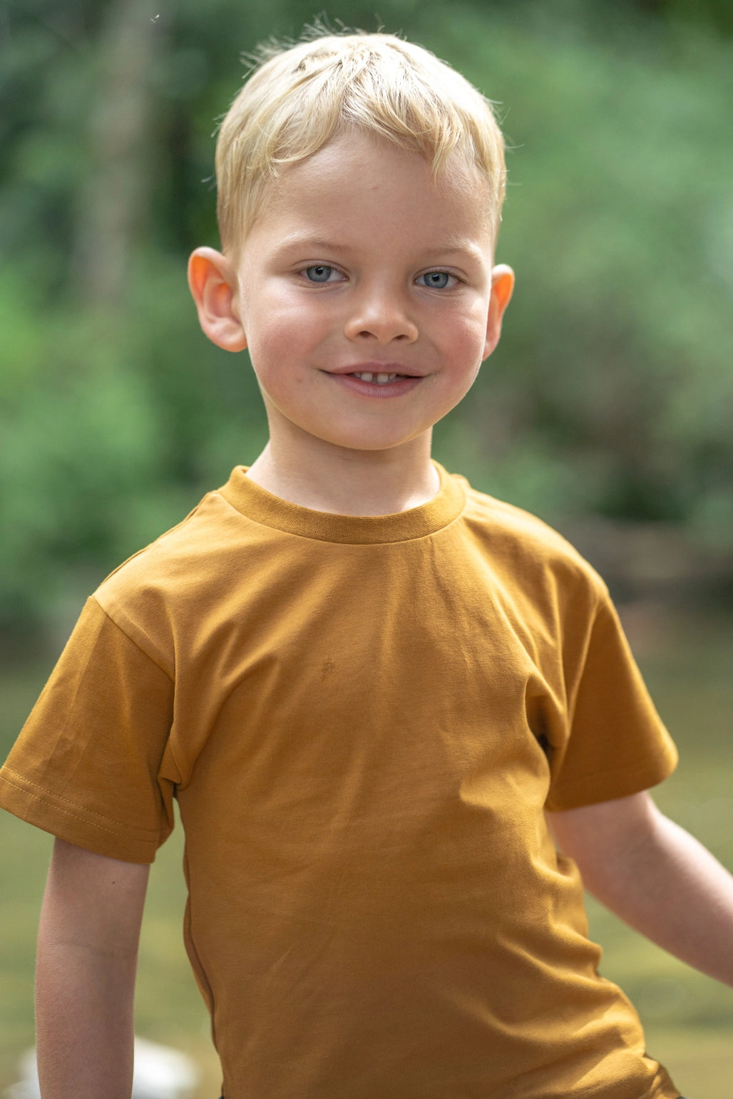 A young blond boy smiling at the camera, wearing a short-sleeve crewneck t-shirt in a warm mustard yellow color, set against a soft, blurred green outdoor background.