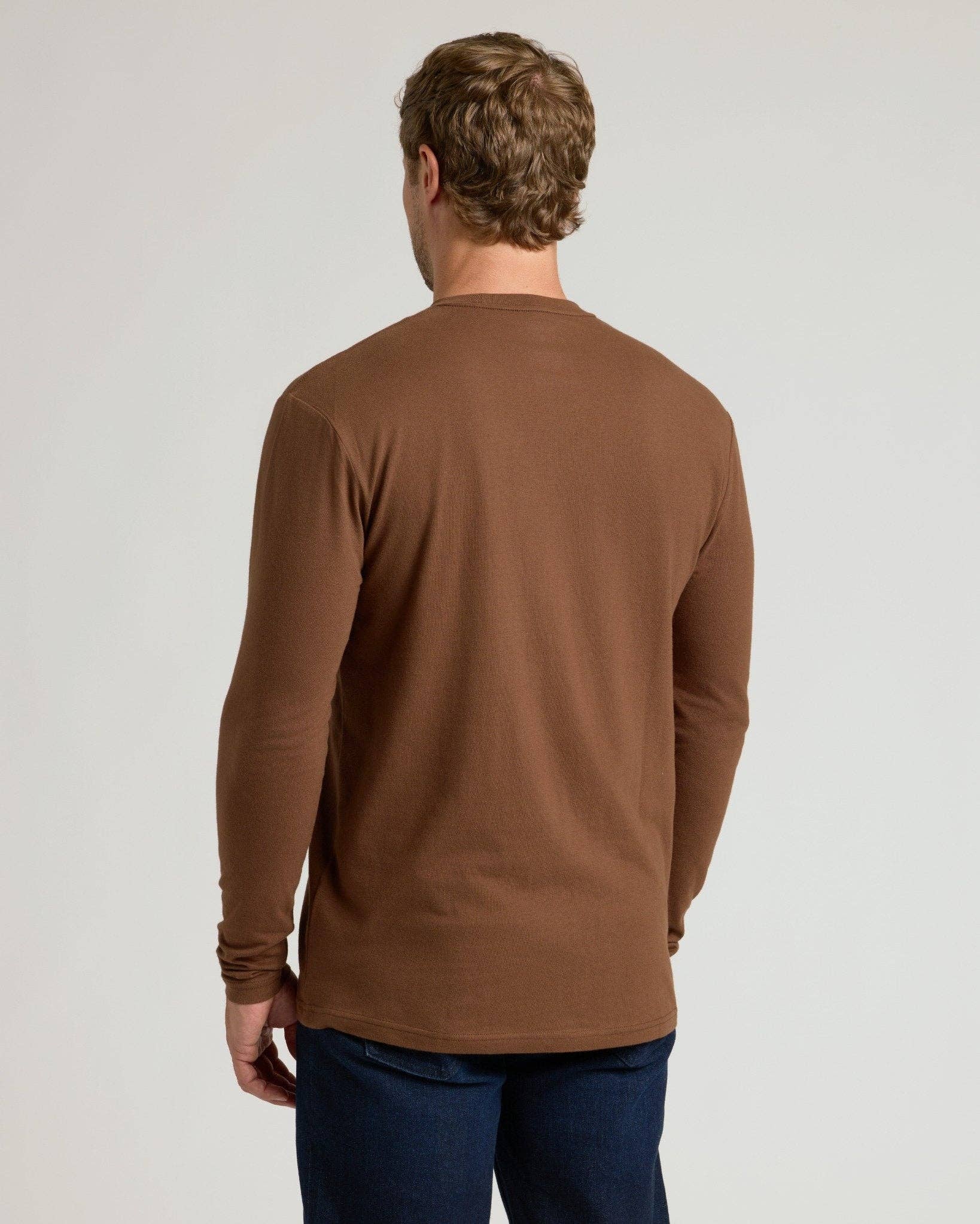 A man viewed from behind wearing a brown long-sleeve crewneck shirt and dark blue jeans, standing against a light gray studio background.