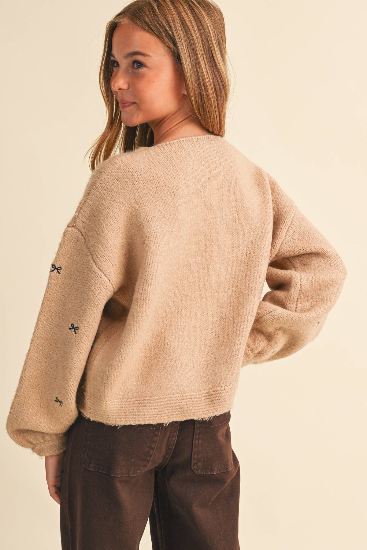 A woman wearing a fuzzy beige knit sweater with small black bow details on the sleeves, shown from a side-back angle against a plain cream background.