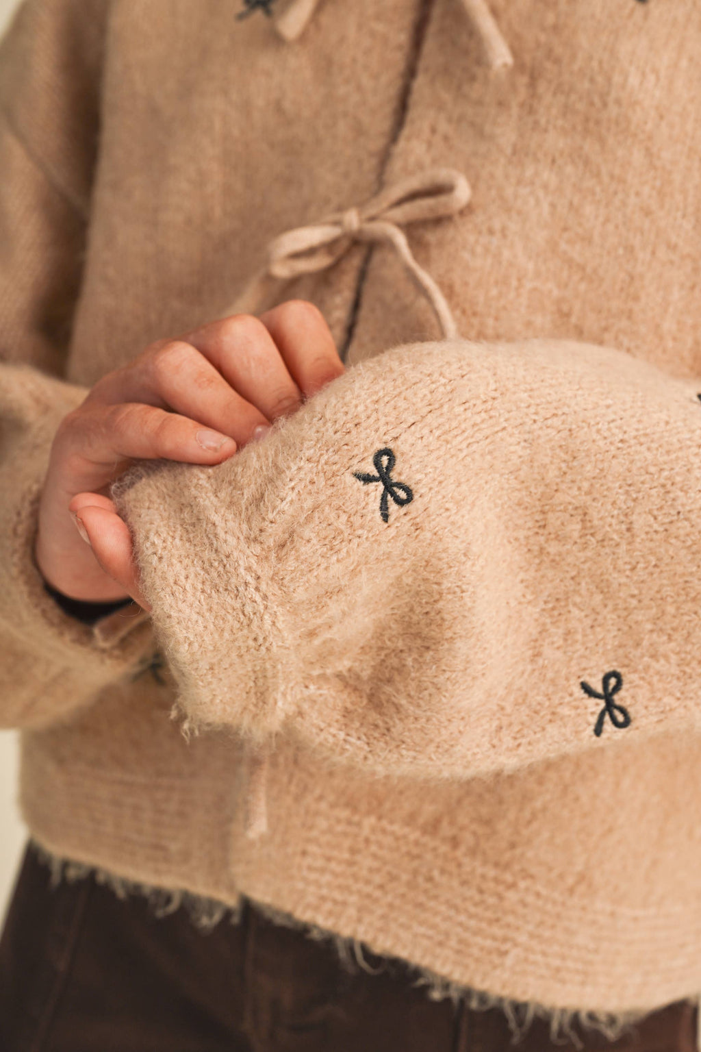Close-up of a person wearing a fuzzy beige knit sweater with small black bow embroidery and a drawstring closure, holding the sleeve to show texture.