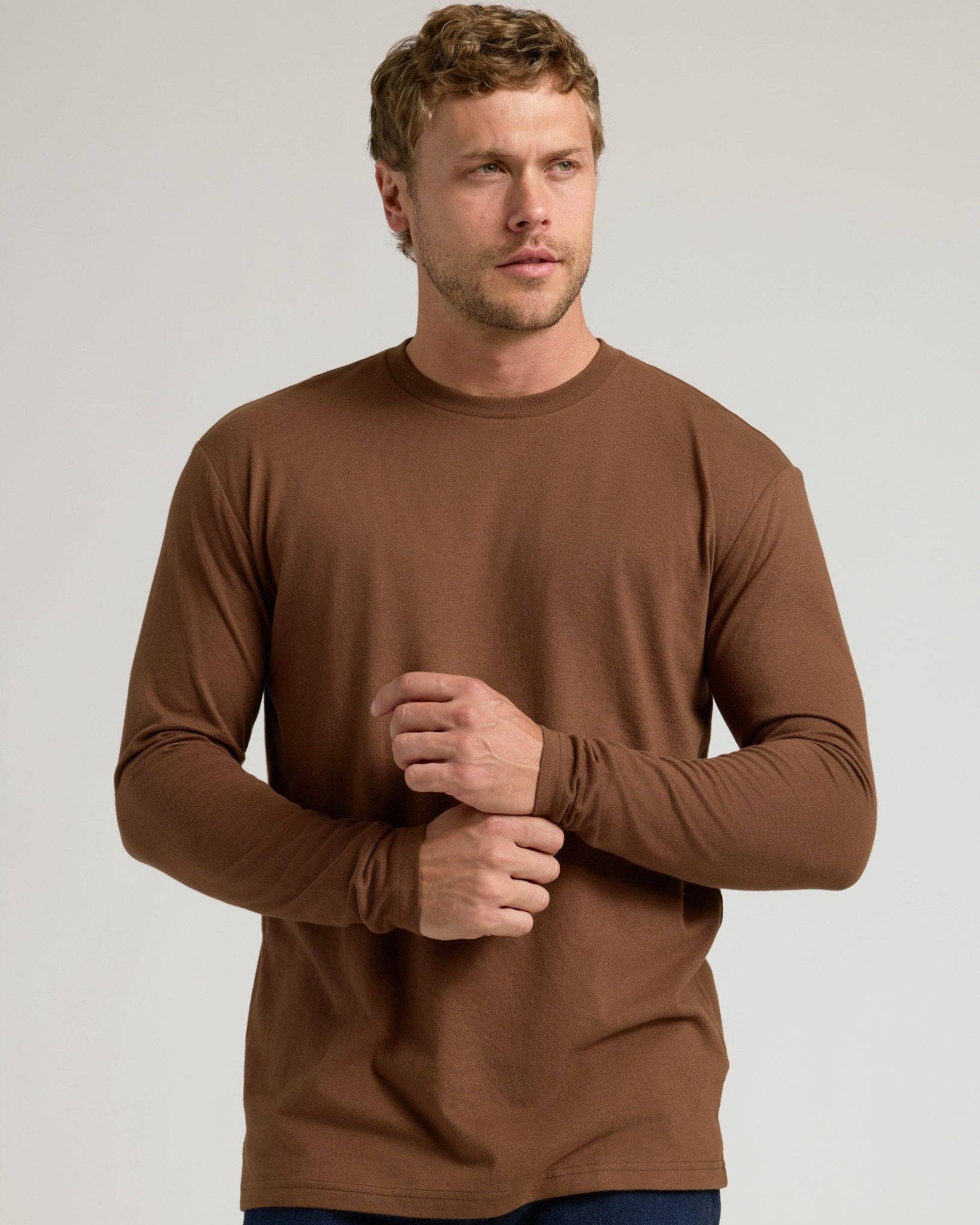 A man wearing a solid brown long-sleeve crewneck shirt, shown from the waist up against a light gray studio background.