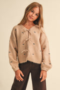 A young girl with long blonde hair wearing a fuzzy beige cropped cardigan with small black bow details and dark brown pants, posing against a solid light beige background.