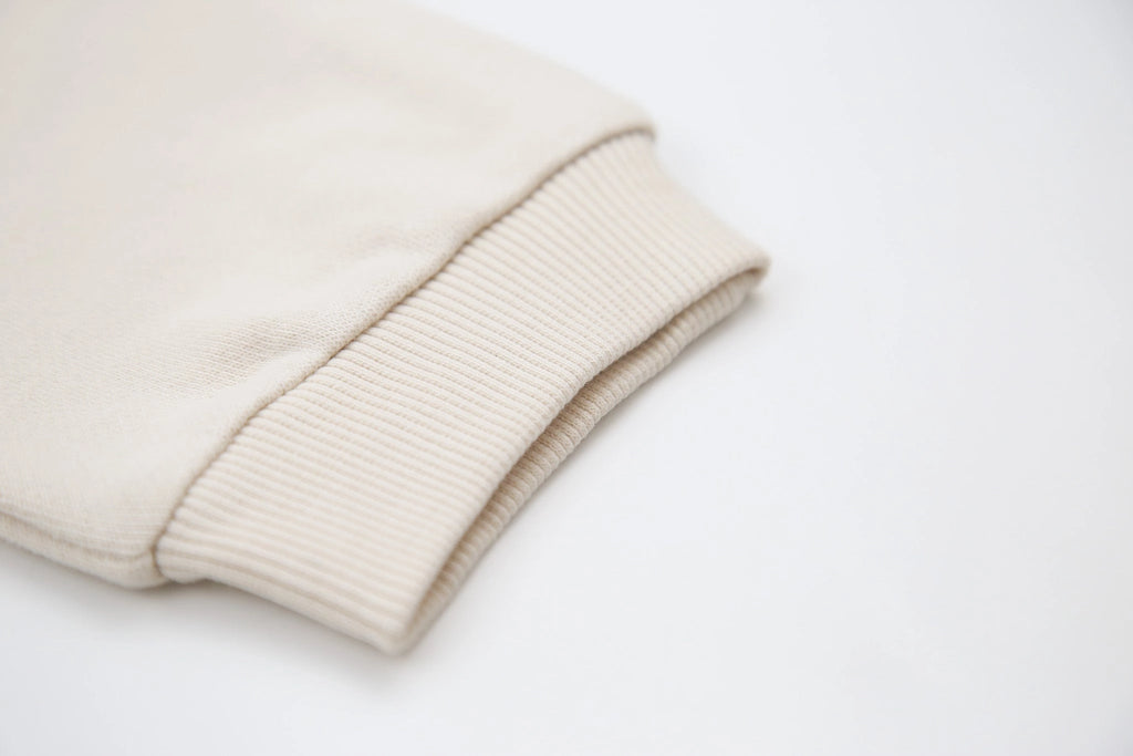 Close-up of a folded ribbed knit garment in a light cream or oatmeal color on a plain white background.
