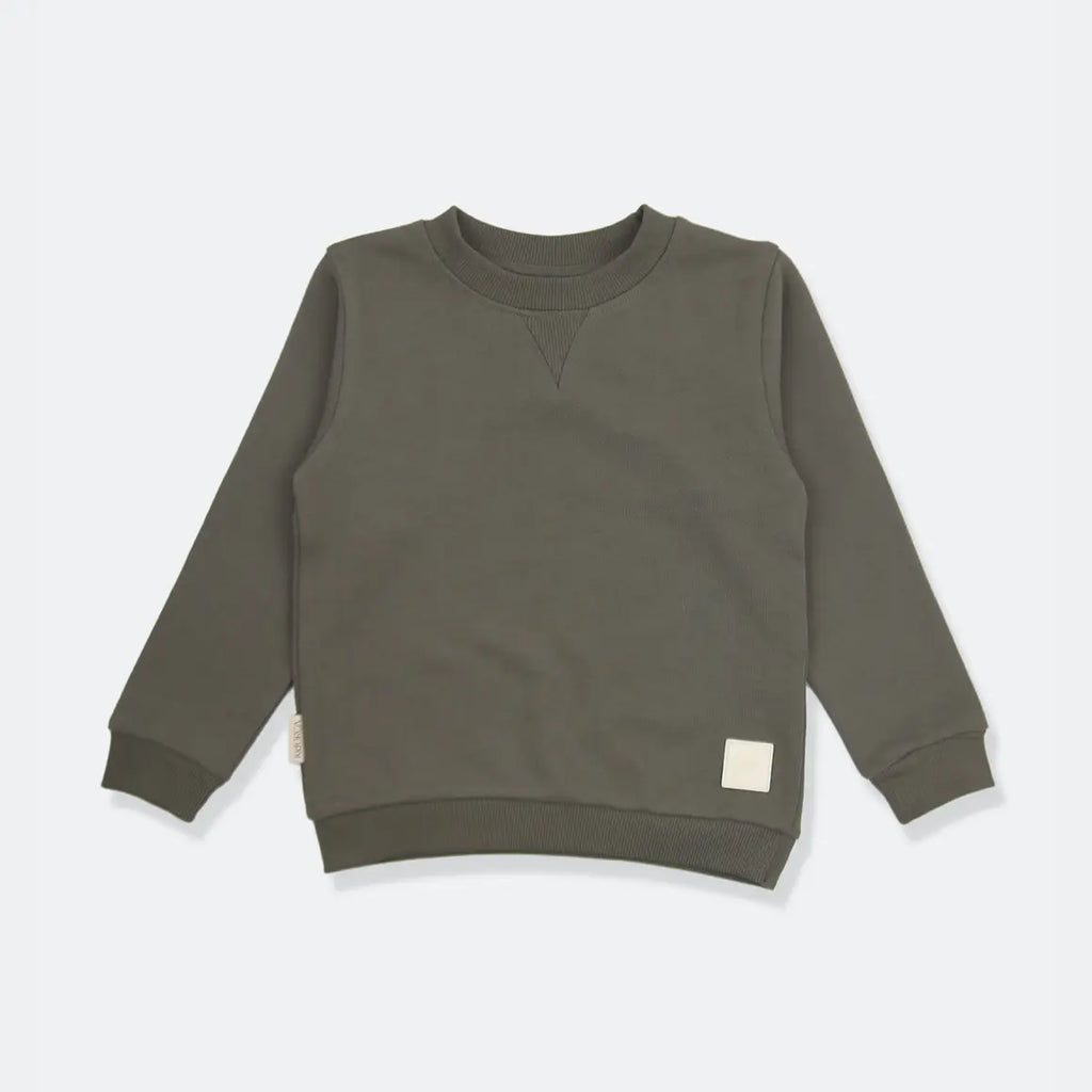 An olive green long-sleeve crewneck sweatshirt for a toddler, laid flat on a light gray background. It features a small cream-colored square patch on the lower right hem and a small fabric tag on the lower left sleeve.